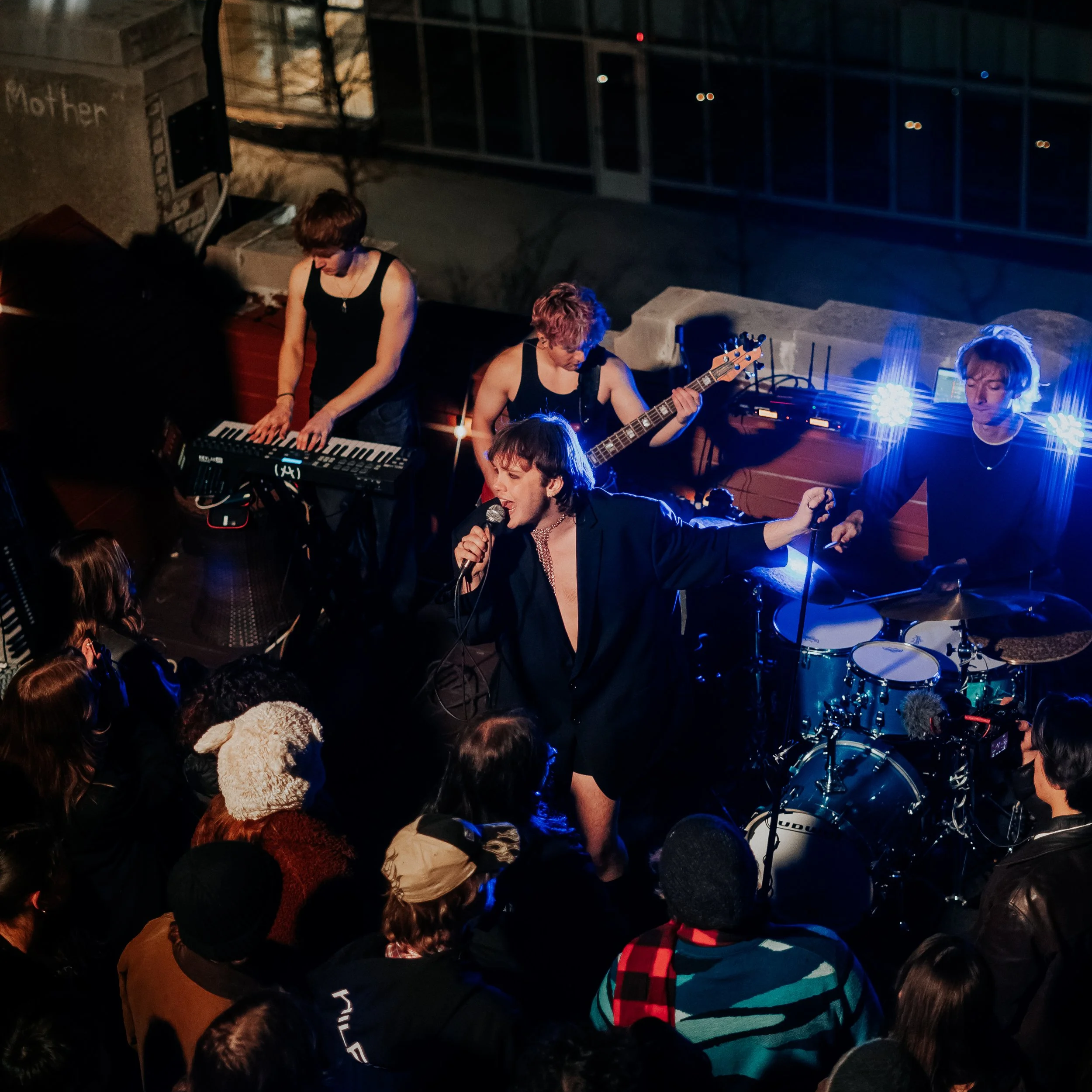 A band performing on stage at night, with a crowd of people watching. The band members include a person playing keyboard, a person playing guitar, a lead singer, and a drummer. The scene is illuminated by colorful stage lights.