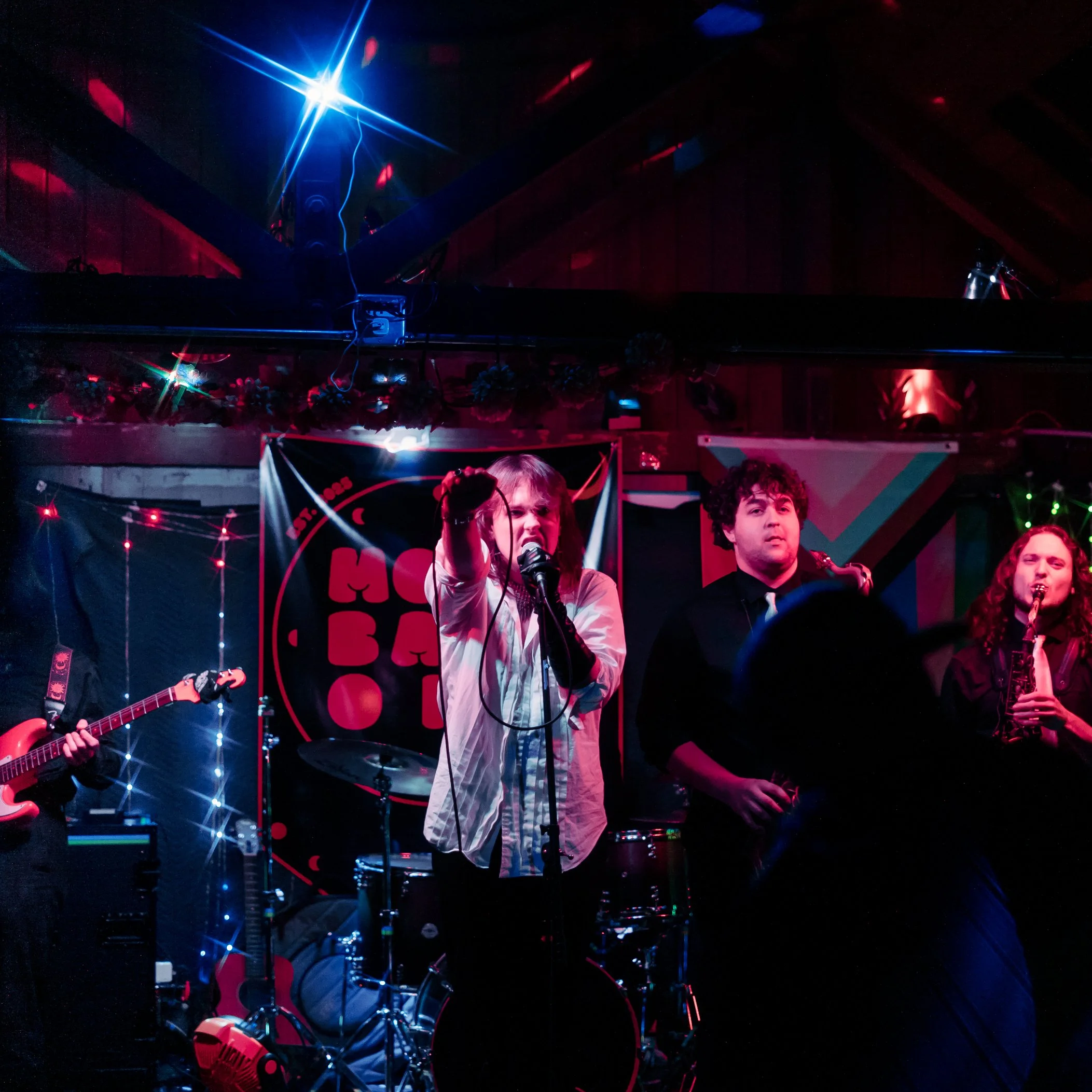 A group of people on stage performing with a band, decorated with string lights and a backdrop with a red logo or text, under stage lighting in a dark venue.