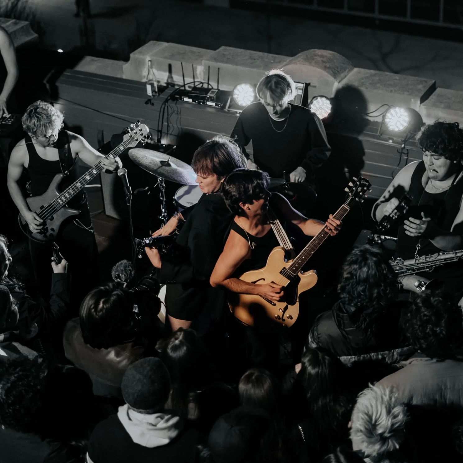 Live music performance with band members playing guitar, drums, bass, and saxophone on stage, with a crowd watching.