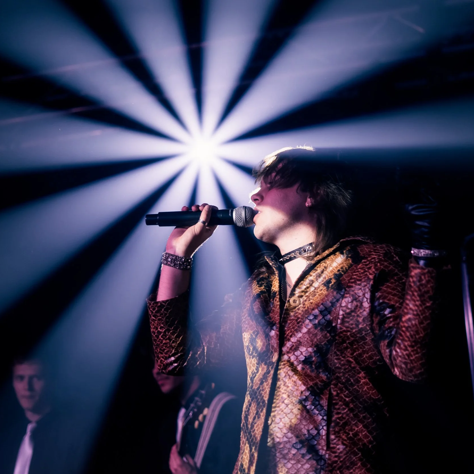 A person singing into a microphone on stage with dramatic lighting and a blue and purple color scheme.
