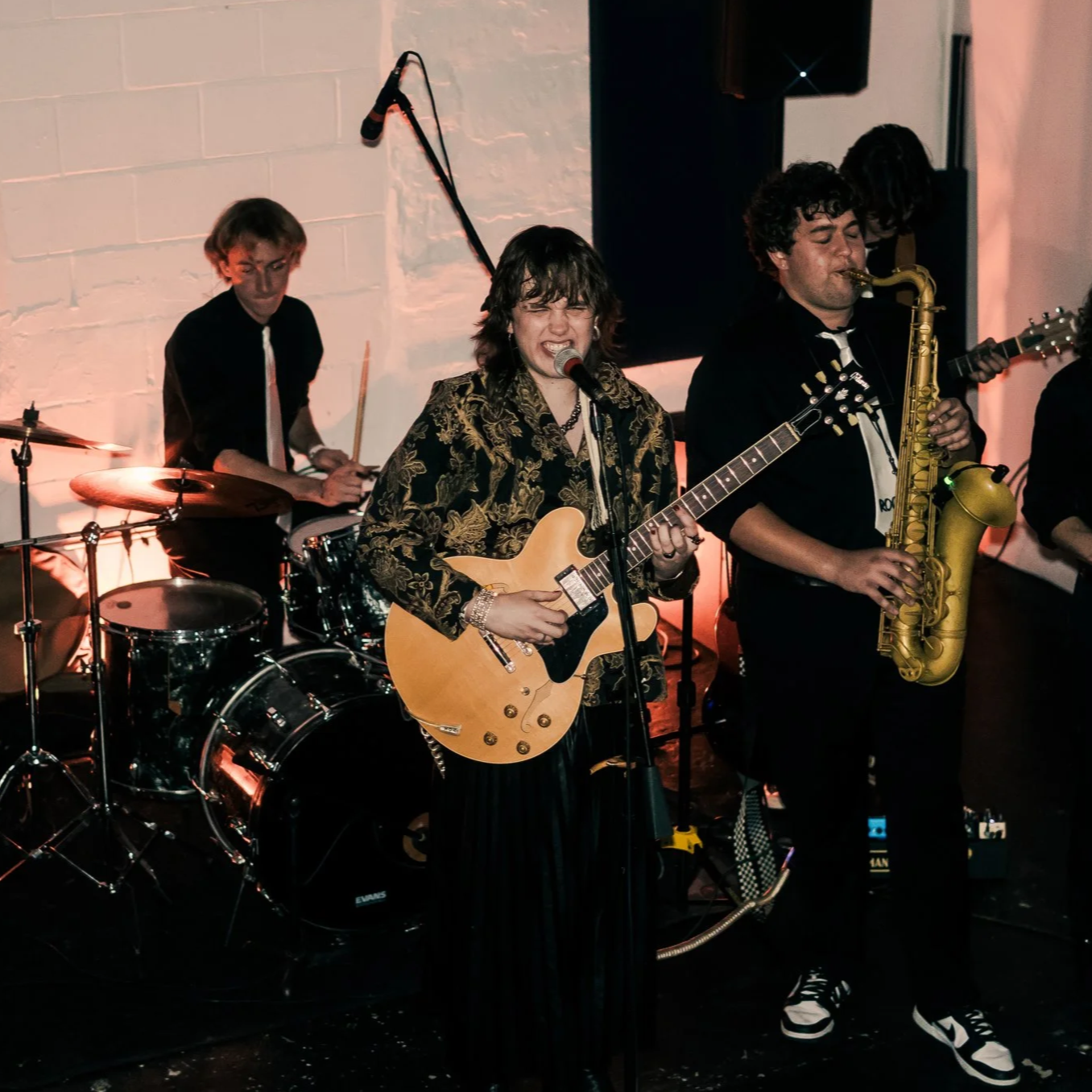 A band performing on stage with a drummer, a guitarist singing into a microphone, and a saxophonist playing. The background features a white brick wall.