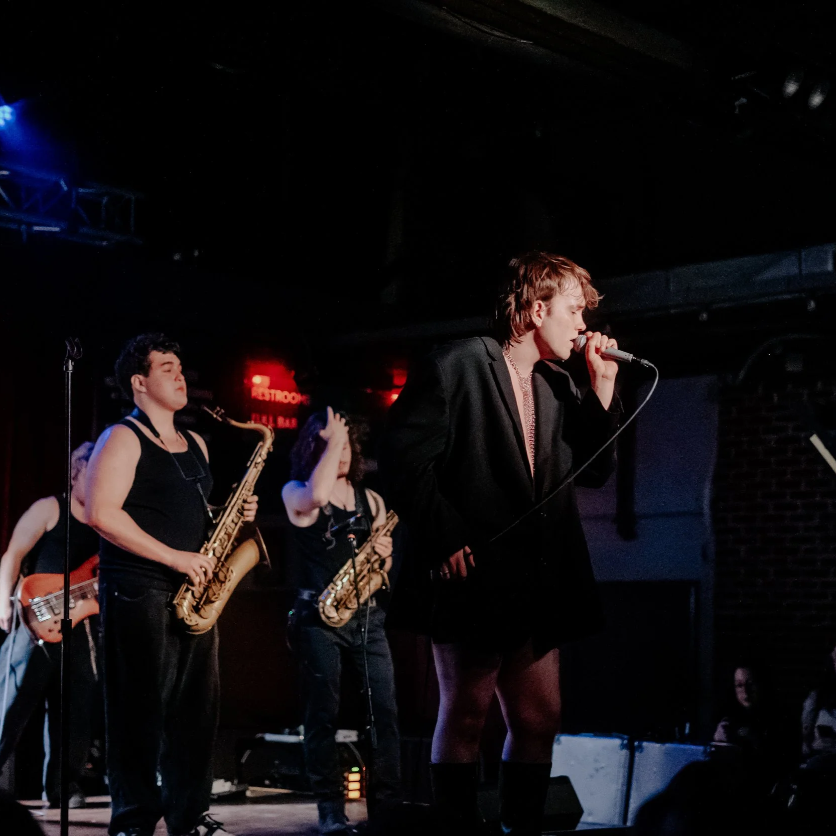 Musicians performing on stage at a nightclub, with a female saxophonist, a singer, and a bass player, under colorful lighting.
