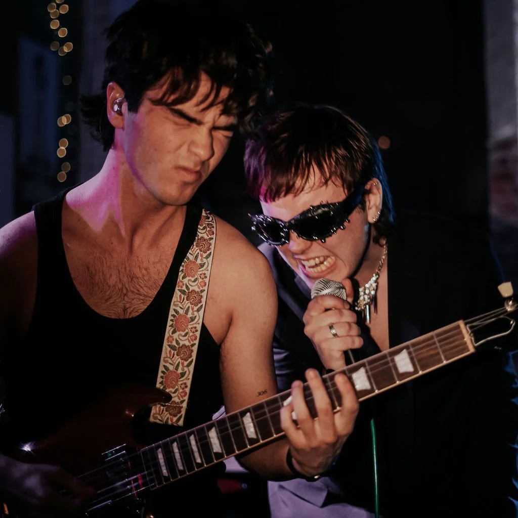 Two musicians perform on stage, with one playing the guitar and the other singing into a microphone, both expressing intense emotion.