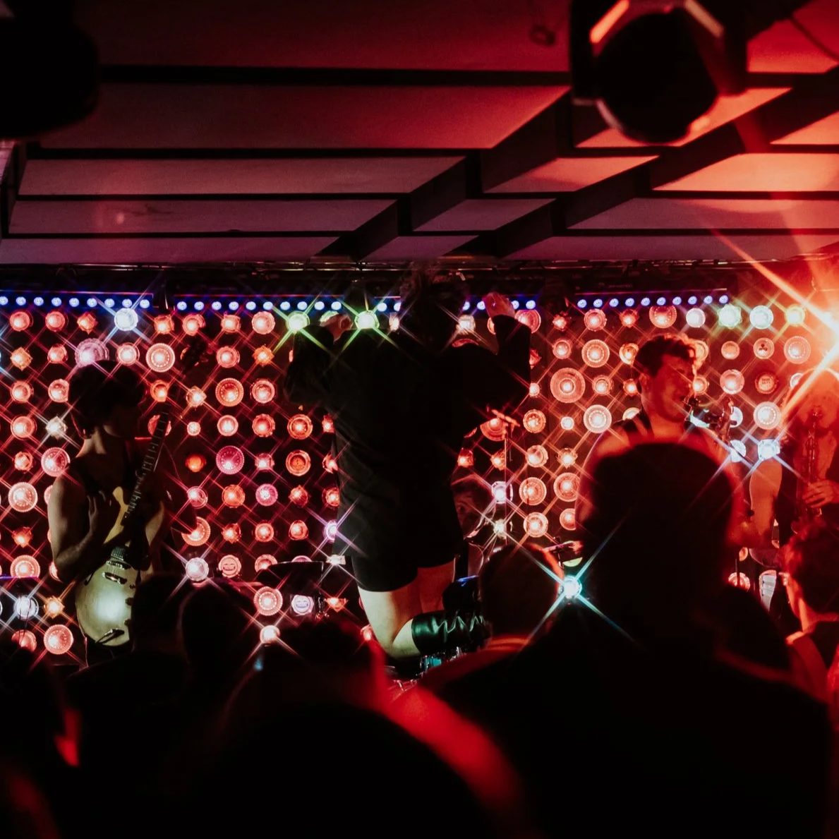 A band performing live on stage with colorful LED lights in the background, featuring a guitarist and a singer, with audience silhouettes in the foreground.