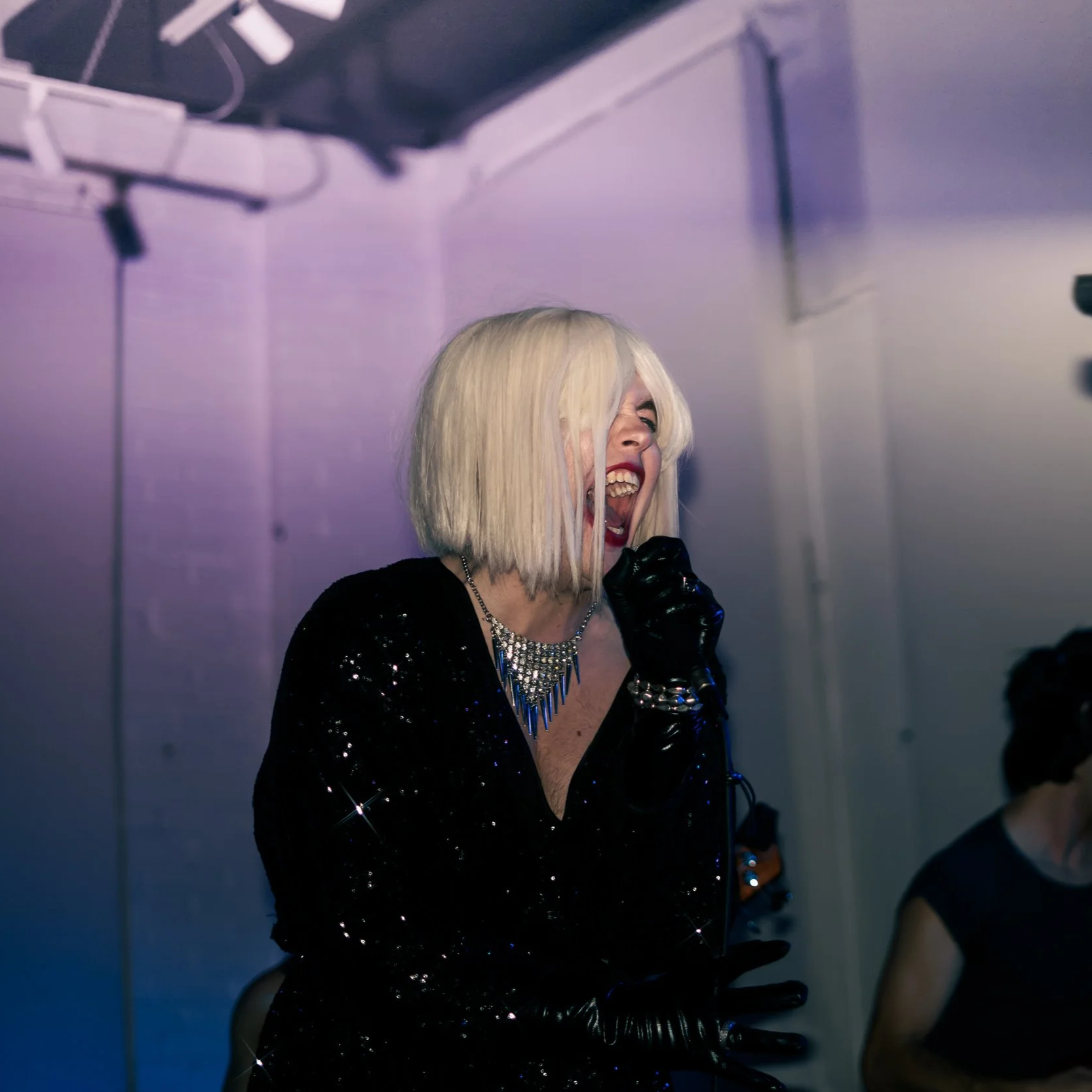 A woman with platinum blonde hair singing into a microphone at a party or performance, wearing a black sequined dress and jewelry.