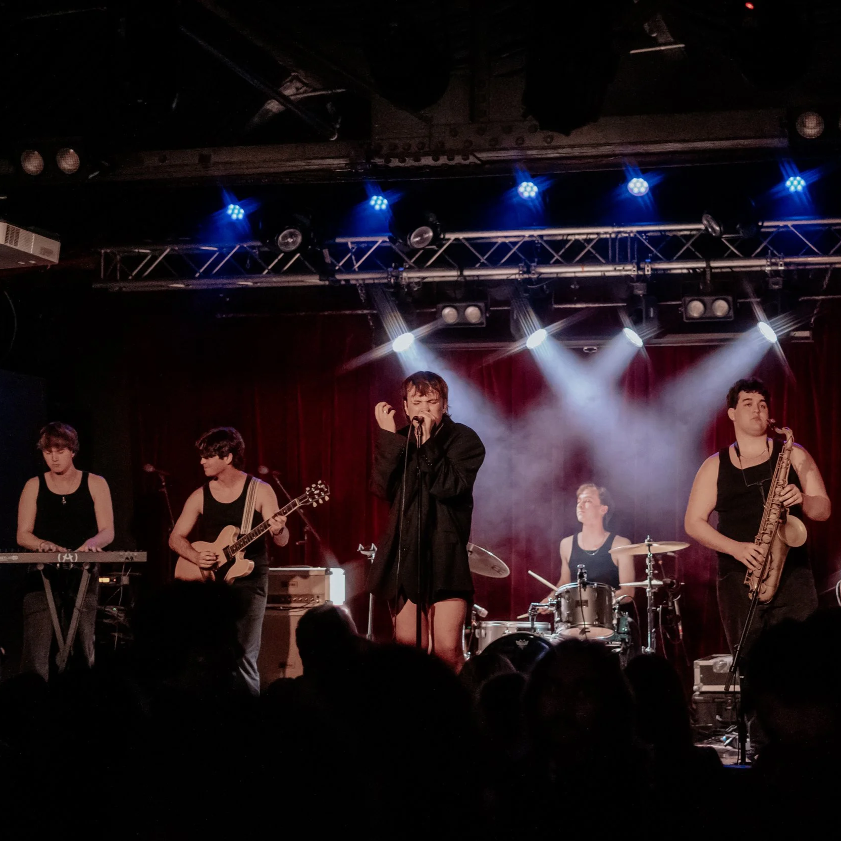A live music performance on stage with a band consisting of a singer, guitarist, saxophonist, and drummer, under colorful stage lighting.