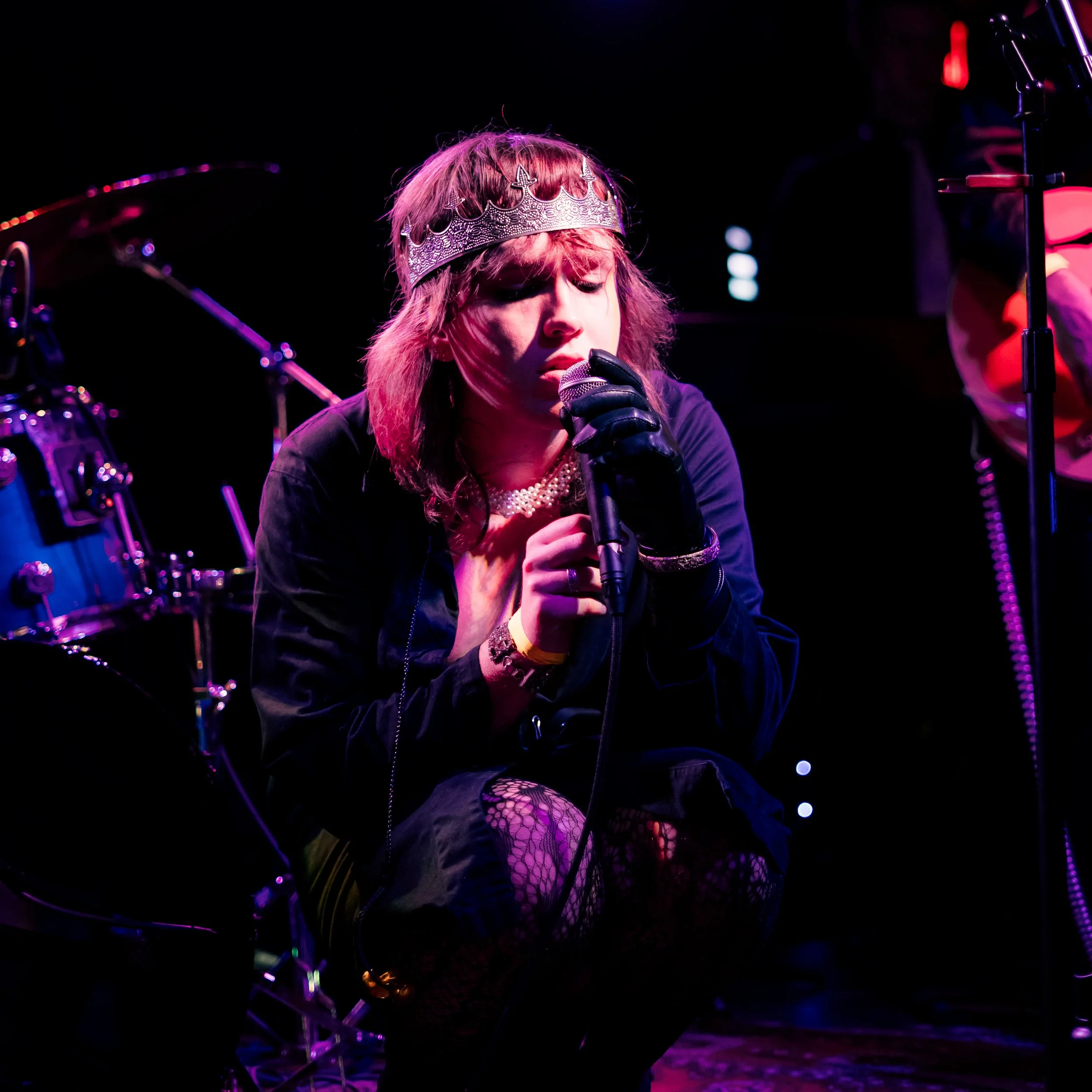 A female performer wearing a crown and fishnet stockings, singing into a microphone on stage with vibrant purple and pink lighting.