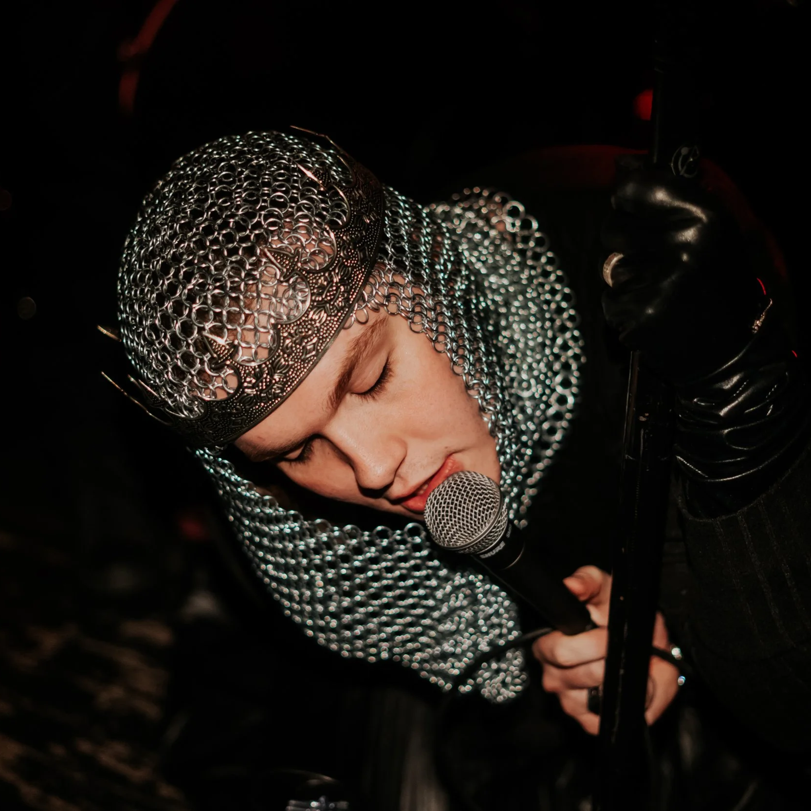 A performer in a shiny chainmail headpiece, singing into a microphone while wearing black gloves, in a dark setting.