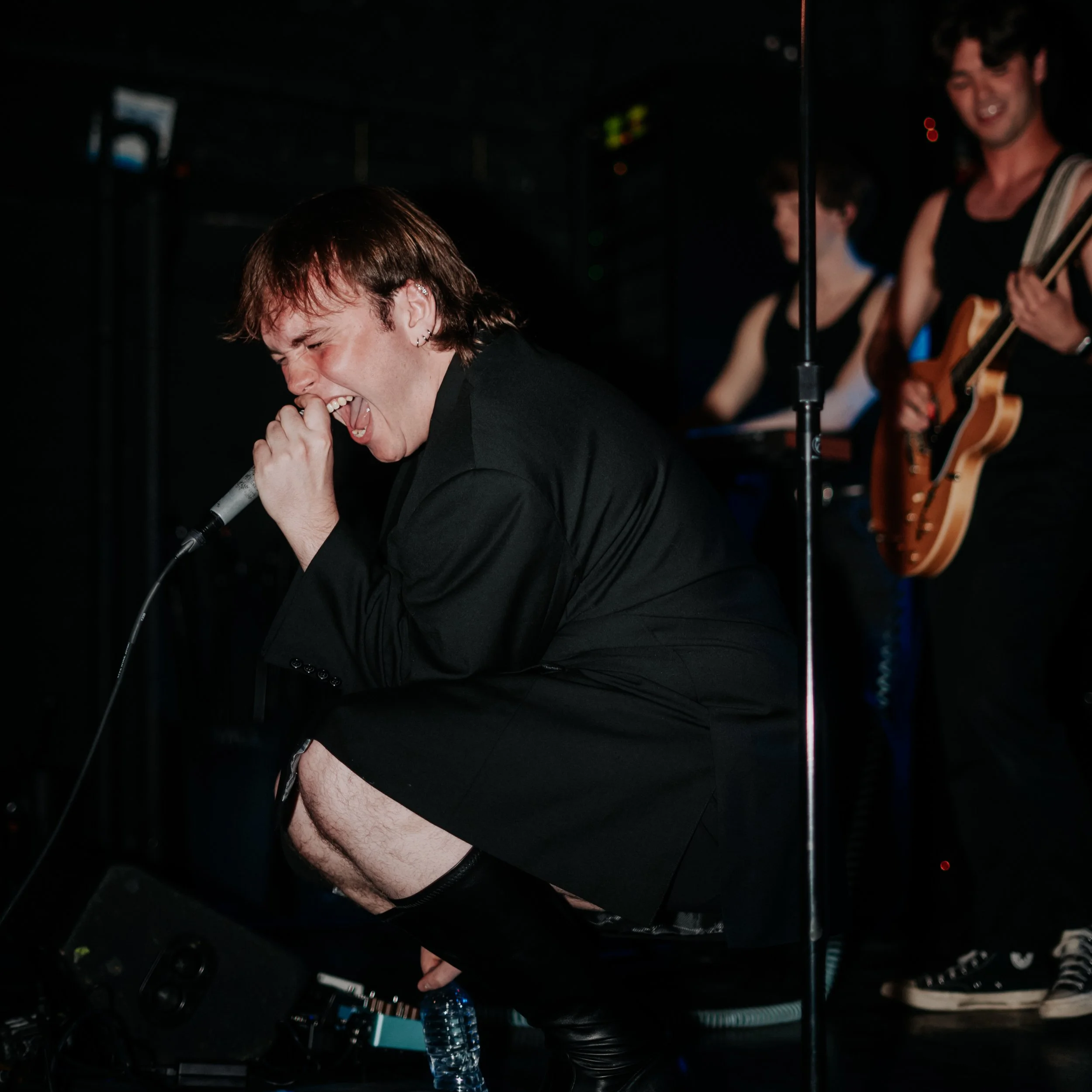 A person with short brown hair singing into a microphone on stage, with another person playing guitar in the background