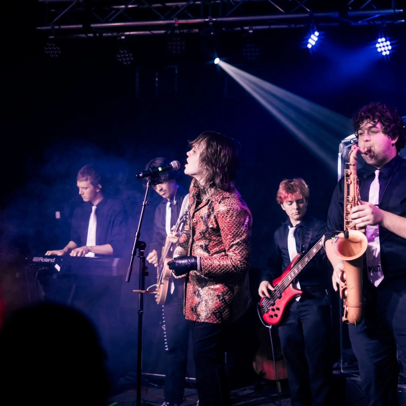 Musicians performing on stage with various instruments, including a keyboard, guitar, and saxophone, under stage lights.