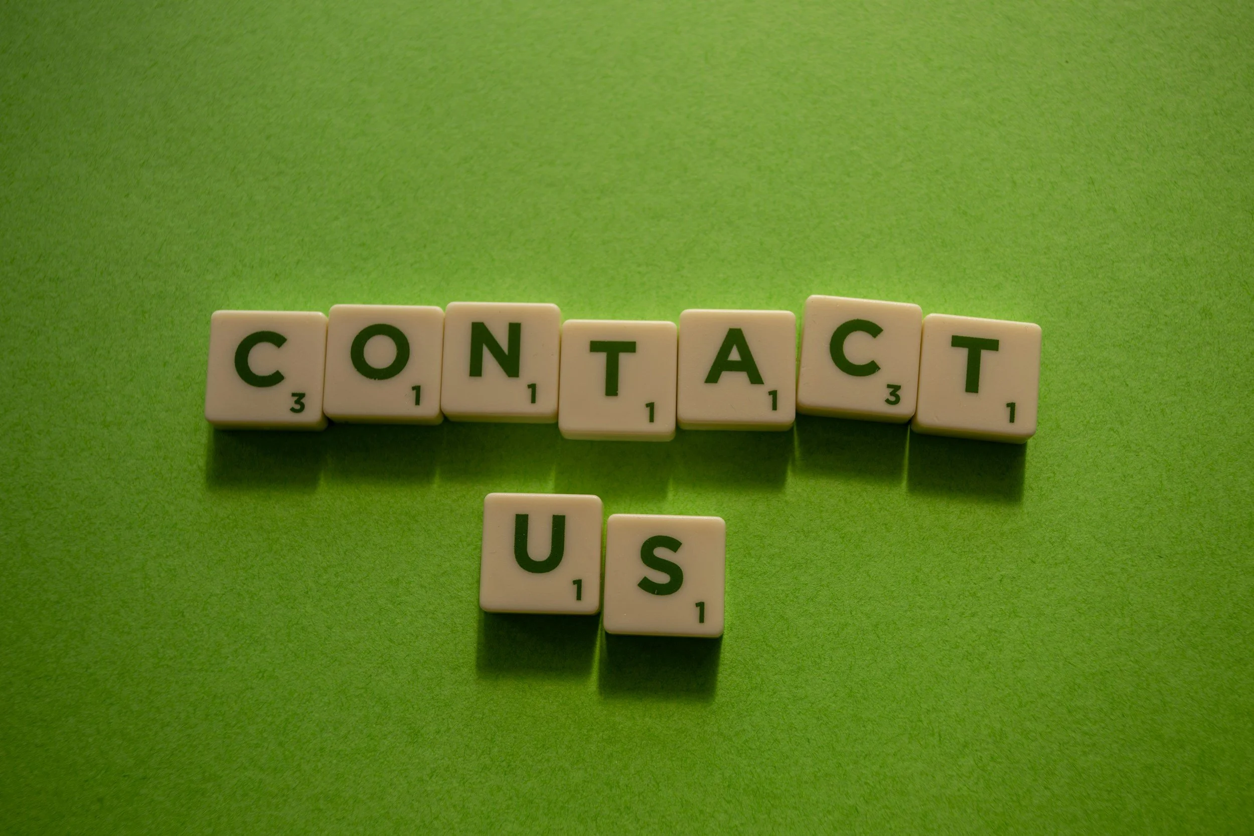 Scrabble tiles spelling out the word "CONTACT US" arranged on a green surface.