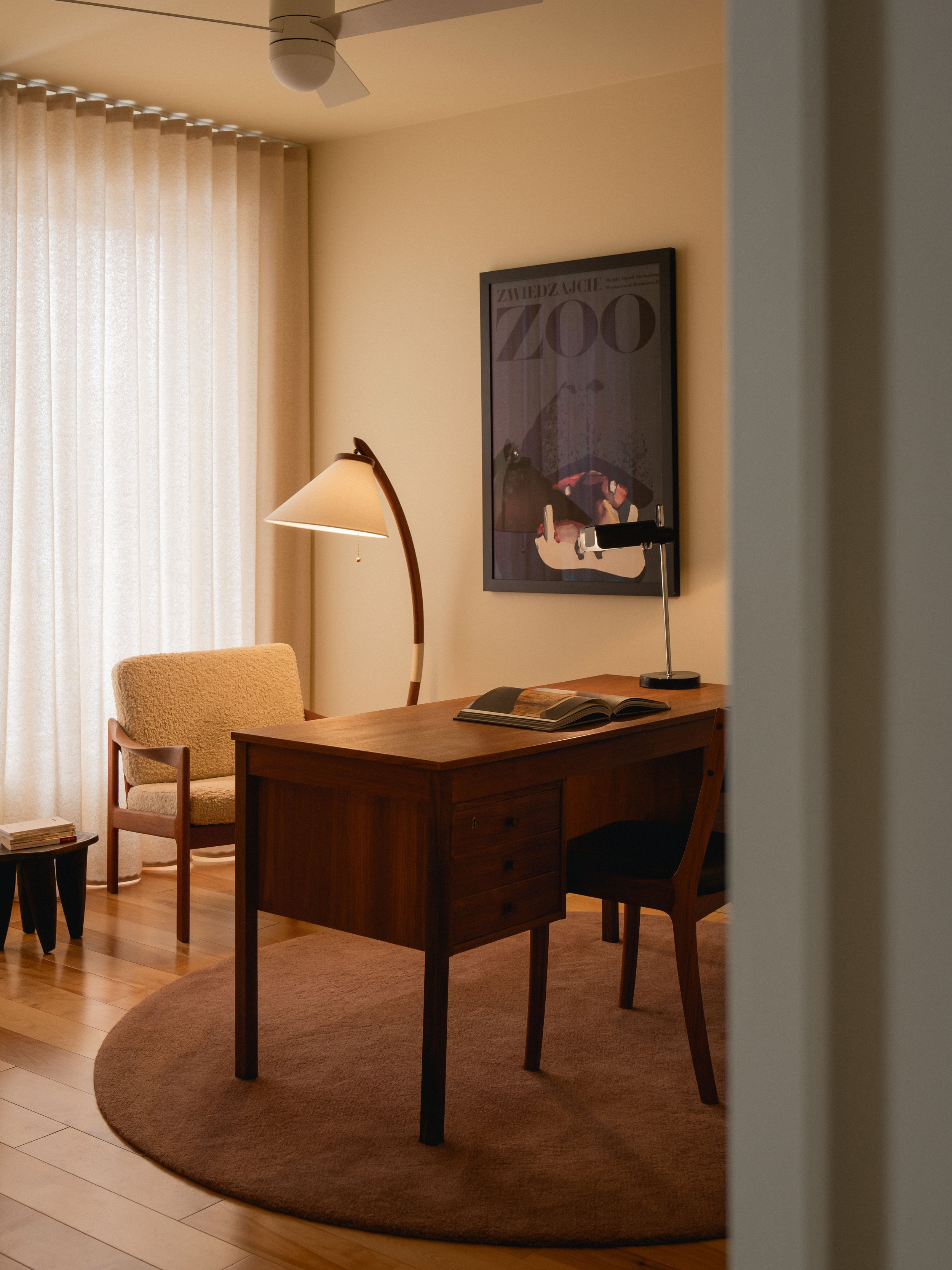 Un salon avec un bureau en bois, une lampe de bureau et une chaise. Une affiche de zoo au mur, un canapé et une petite table. Tapis rond en dessous du bureau.