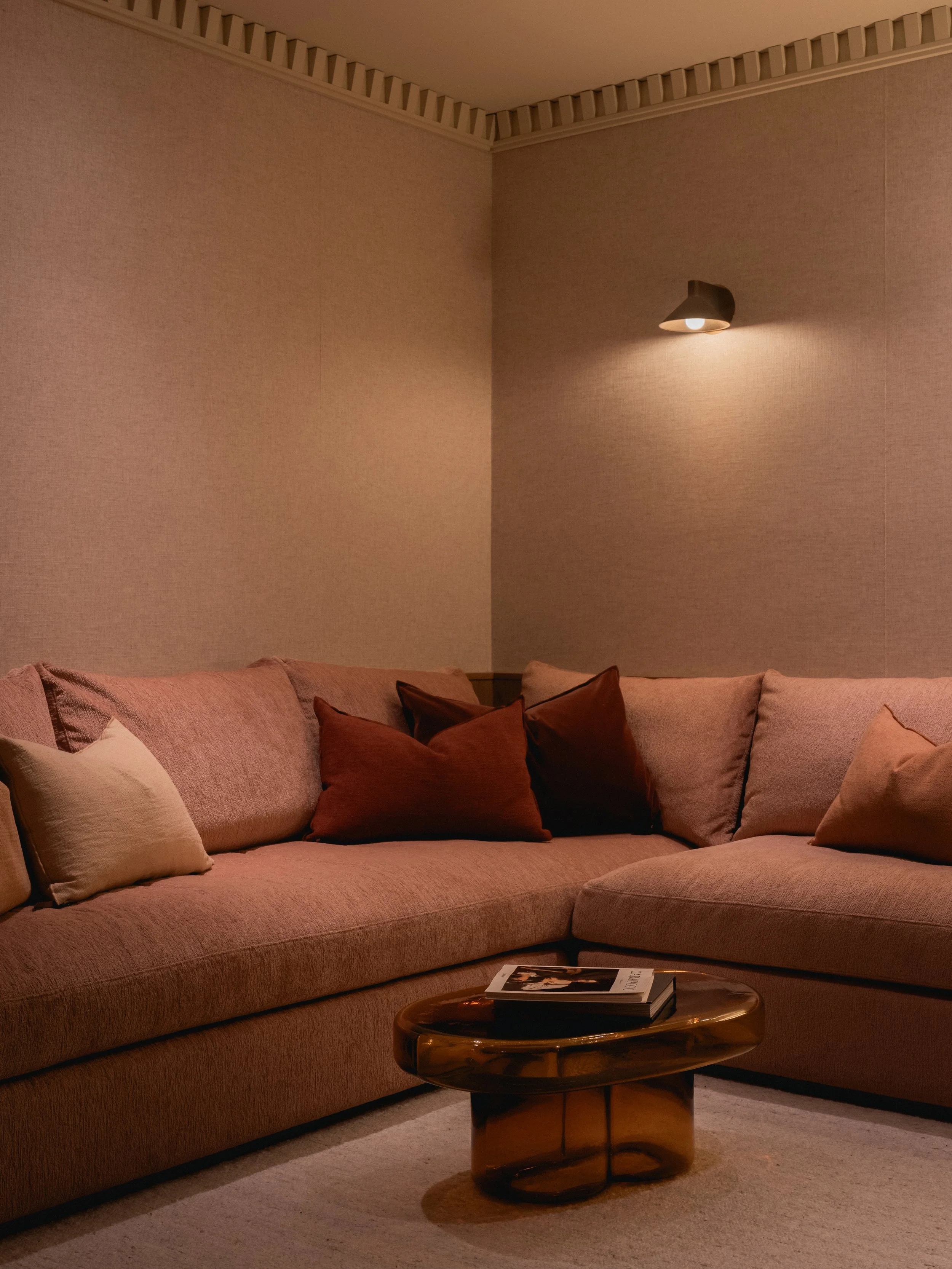 Un salon avec un canapé rose clair, plusieurs coussins de différentes couleurs, une table en bois foncé avec un livre dessus, un mur beige clair et un éclairage d'appoint au mur.