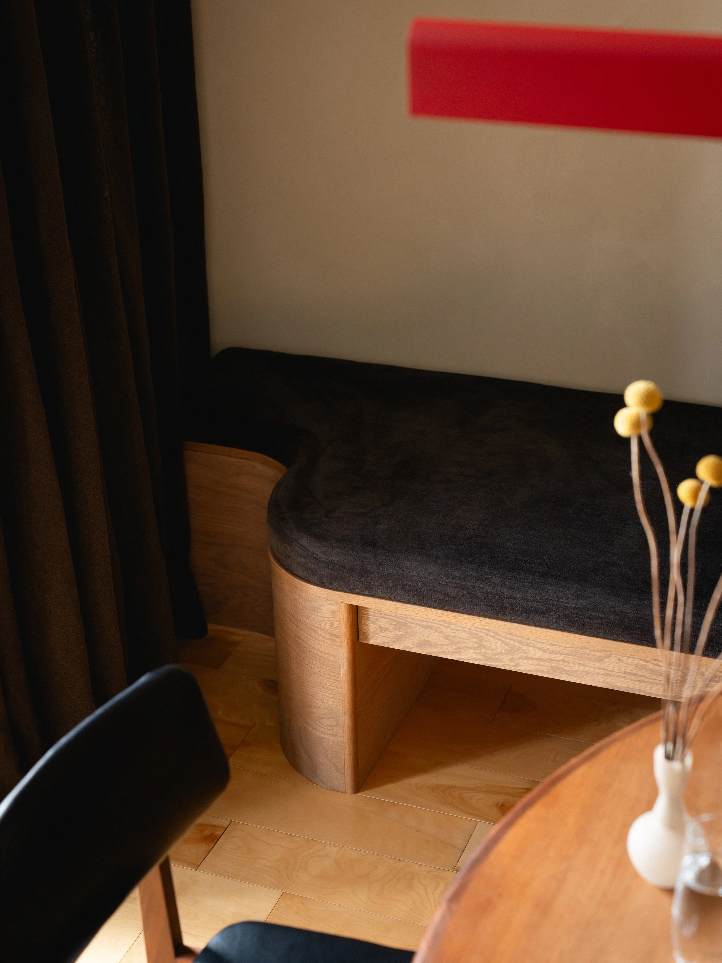 Intérieur d'une pièce avec un banc en bois et un vase contenant des fleurs jaunes, une table ronde en bois clair, un mur beige, une suspension rouge, une chaise noire en avant-plan et des rideaux prunes.