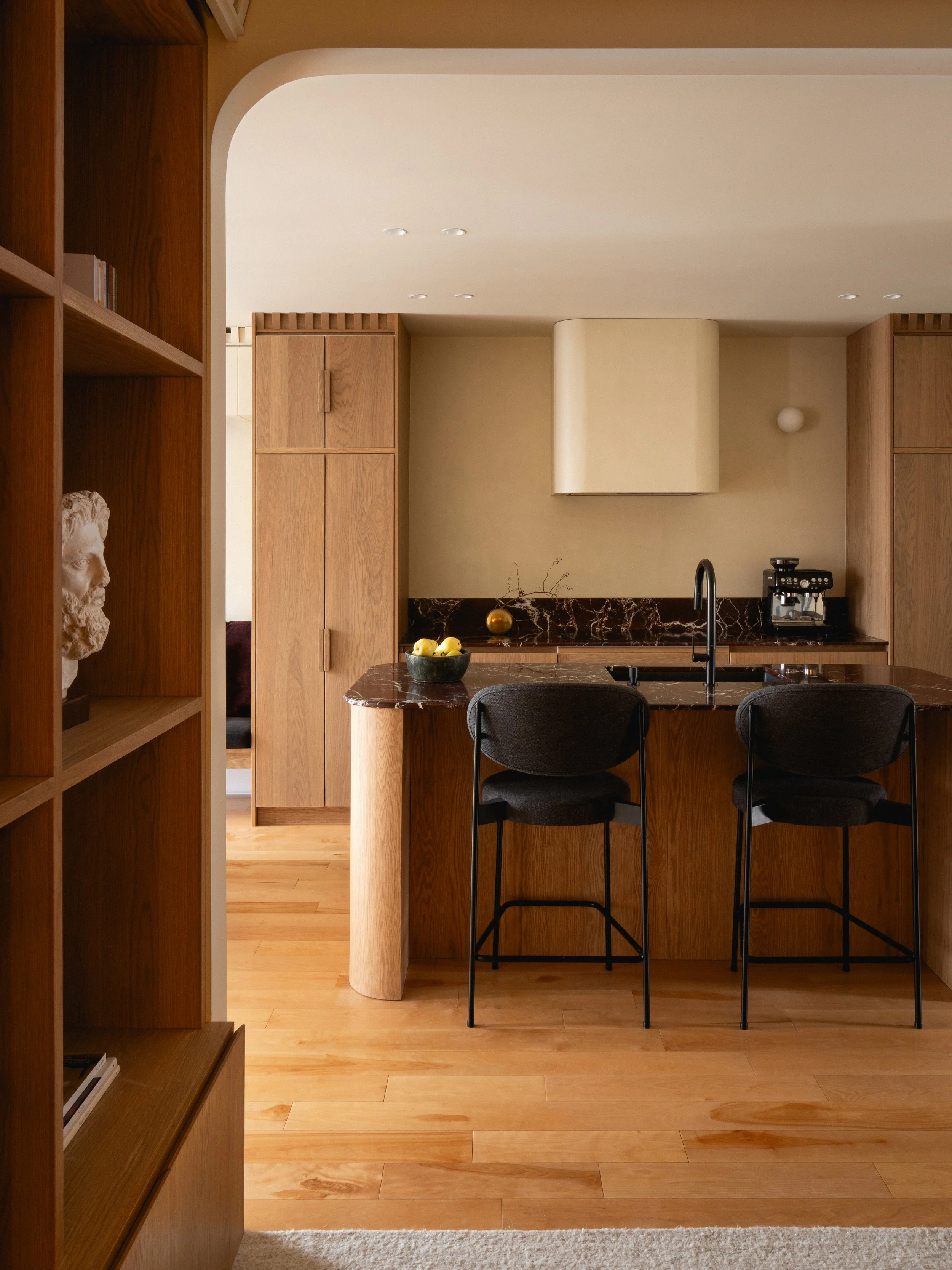 Une cuisine moderne avec un plan de travail en marbre, deux chaises en bois avec assises en tissu noir, des armoires en bois clair, un évier noir, et un mur beige avec un appareil à café noir, un vase avec des branches et des poires dans un bol vert.