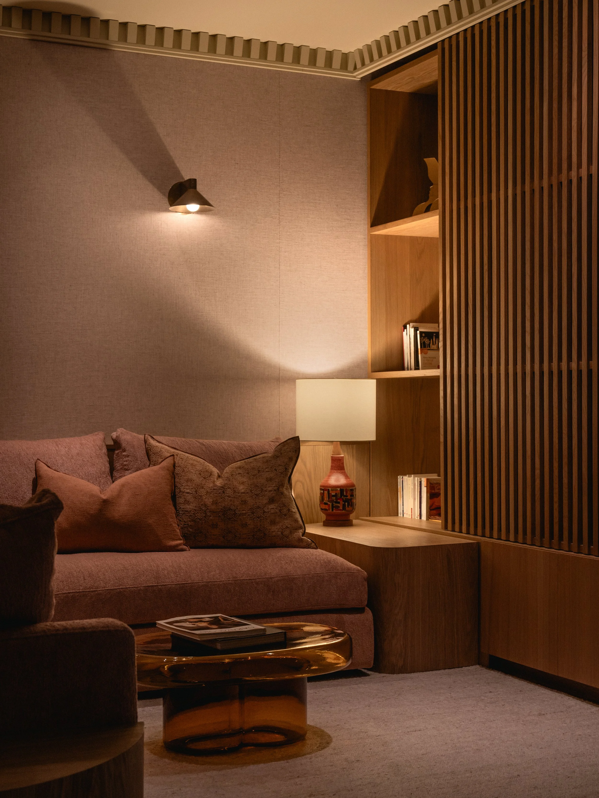 Un coin salon avec un canapé rose, des coussins assortis, une table basse en forme de disque, une lampe de table sur une étagère en bois, des livres et un trophée. Mur de couleur neutre avec une bibliothèque en bois ajouré et une lampe murale.