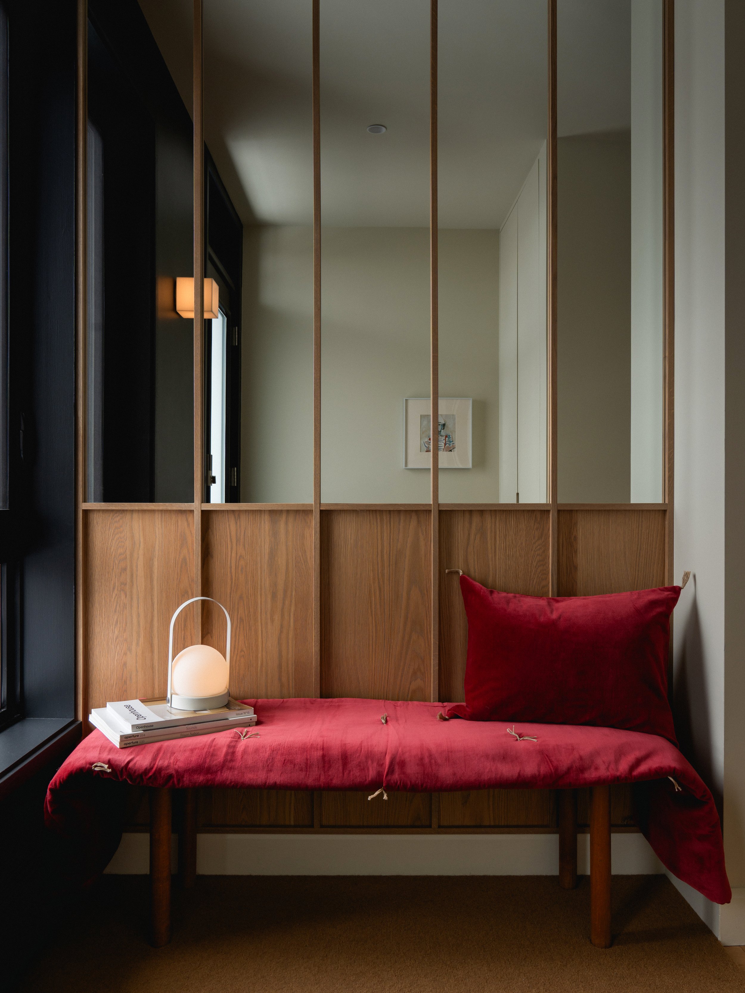 Un banc avec une jetée rouge et un coussin rouge, une lampe blanche sur une pile de livres, un mur blanc avec un tableau et un miroir avec baguettes de bois.