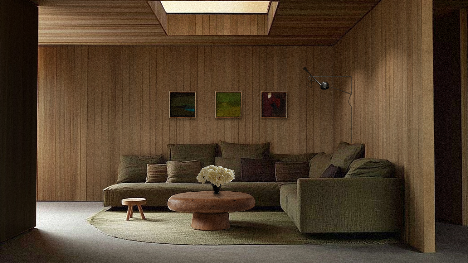 Aménagement intérieur minimaliste avec un canapé vert, un tapis beige, une table en bois avec des fleurs blanches, trois tableaux abstraits sur un mur en bois, et une lampe sur un bras ajustable.