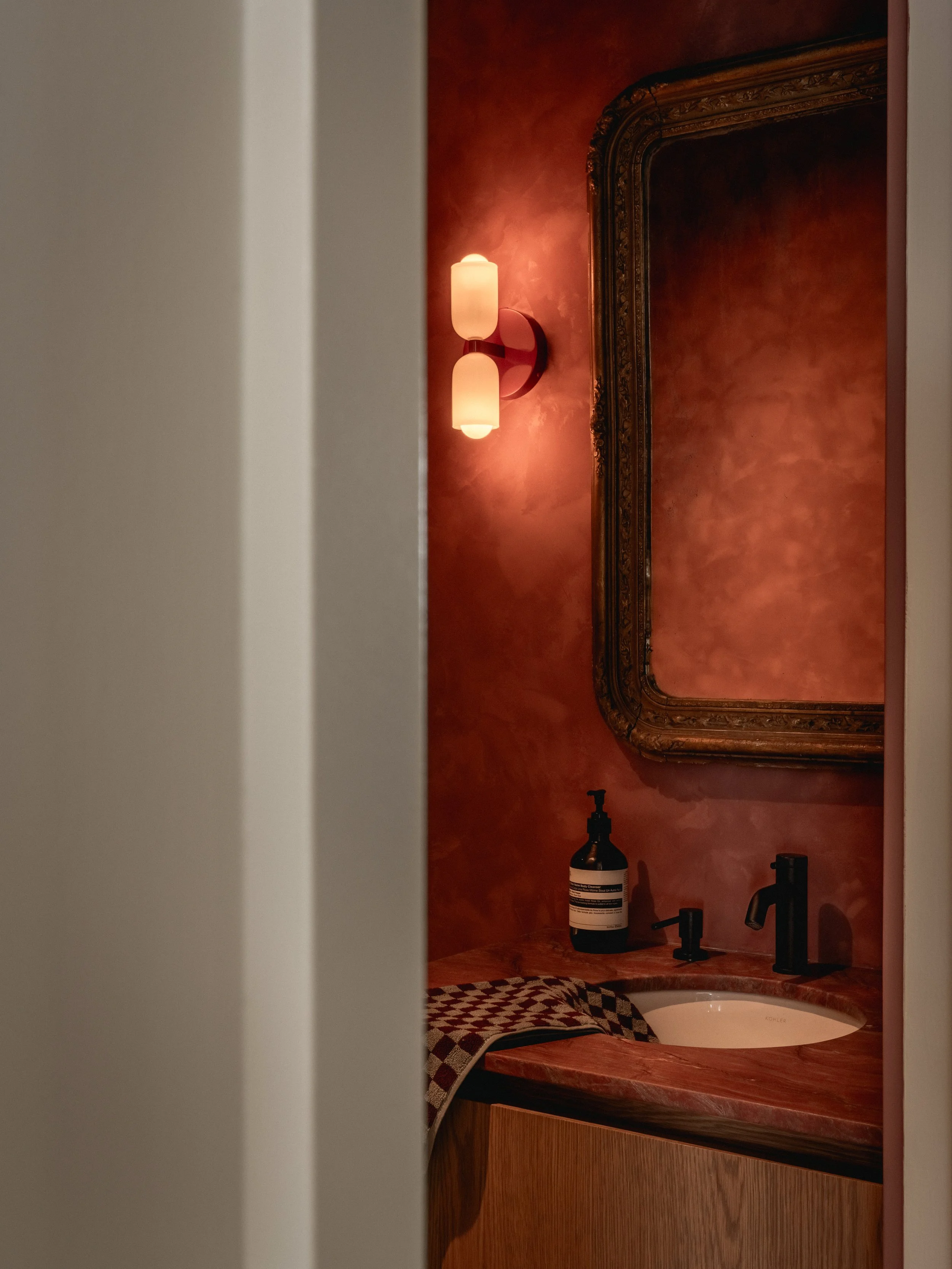 Un miroir encadré en bois au mur rouge, avec une lampe murale et un lavabo. Sur le comptoir, une bouteille de produit de soins et deux robinets.