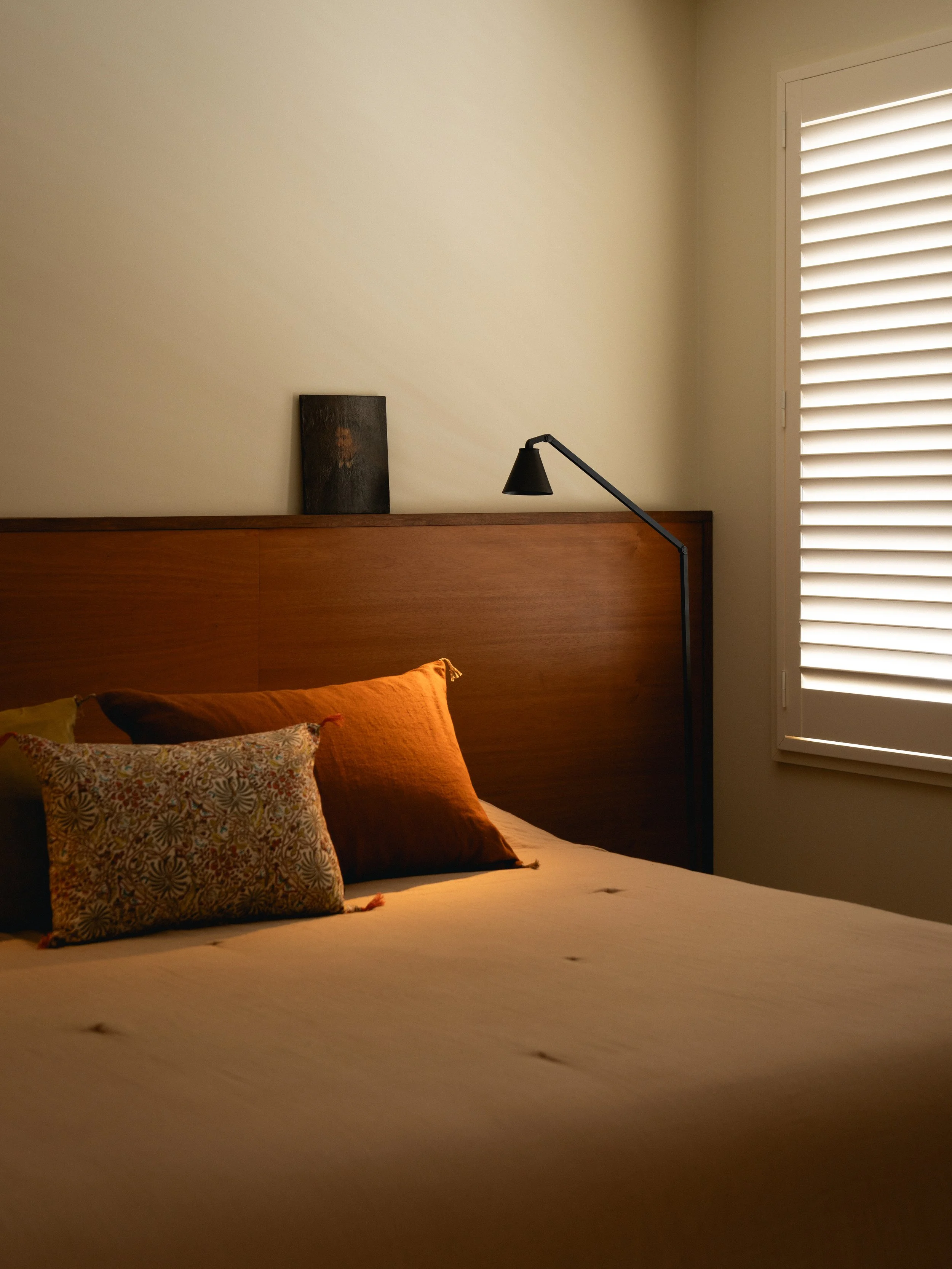 Chambre chaleureuse avec lit, oreillers colorés, lampadaire et fenêtre à volets blancs.
