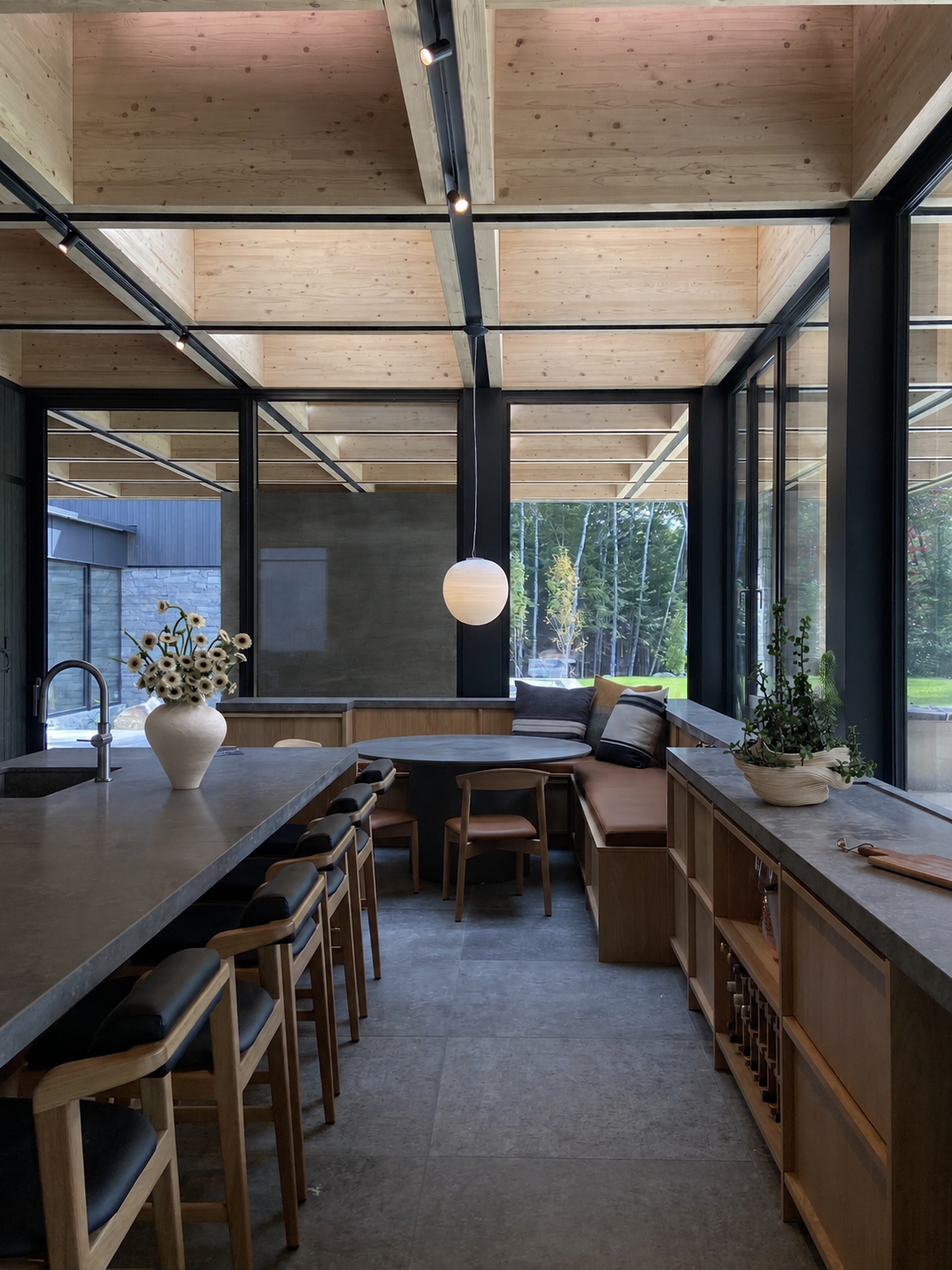 Intérieur d'une cuisine moderne avec des éléments en bois, un comptoir avec un vase de fleurs, un coin repas avec des coussins et des chaises en bois, beaucoup de lumière naturelle et de grandes fenêtres donnant sur un jardin.