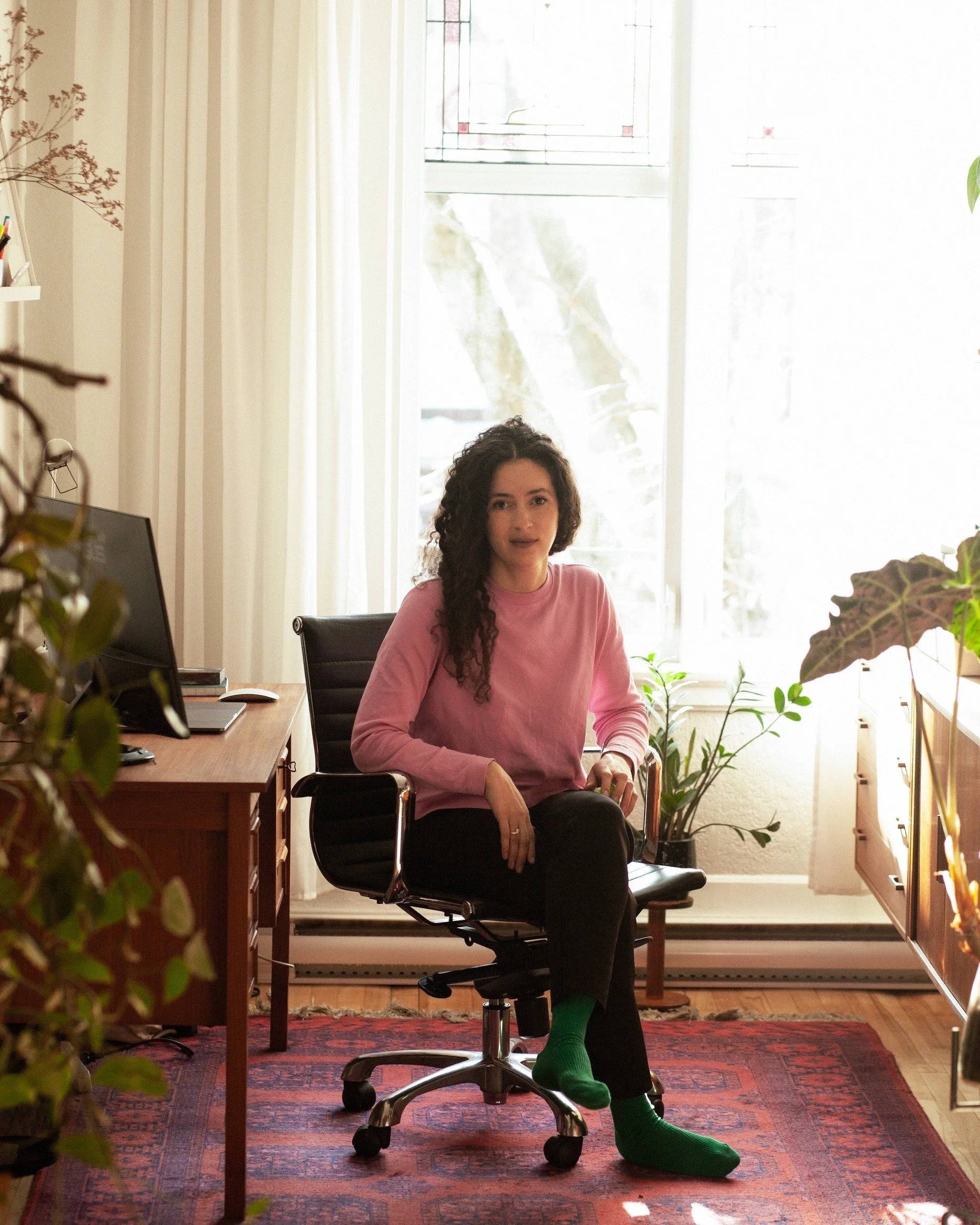 Une femme assise sur une chaise de bureau dans une pièce lumineuse, entourée de plantes et de meubles en bois, regardant vers la caméra.