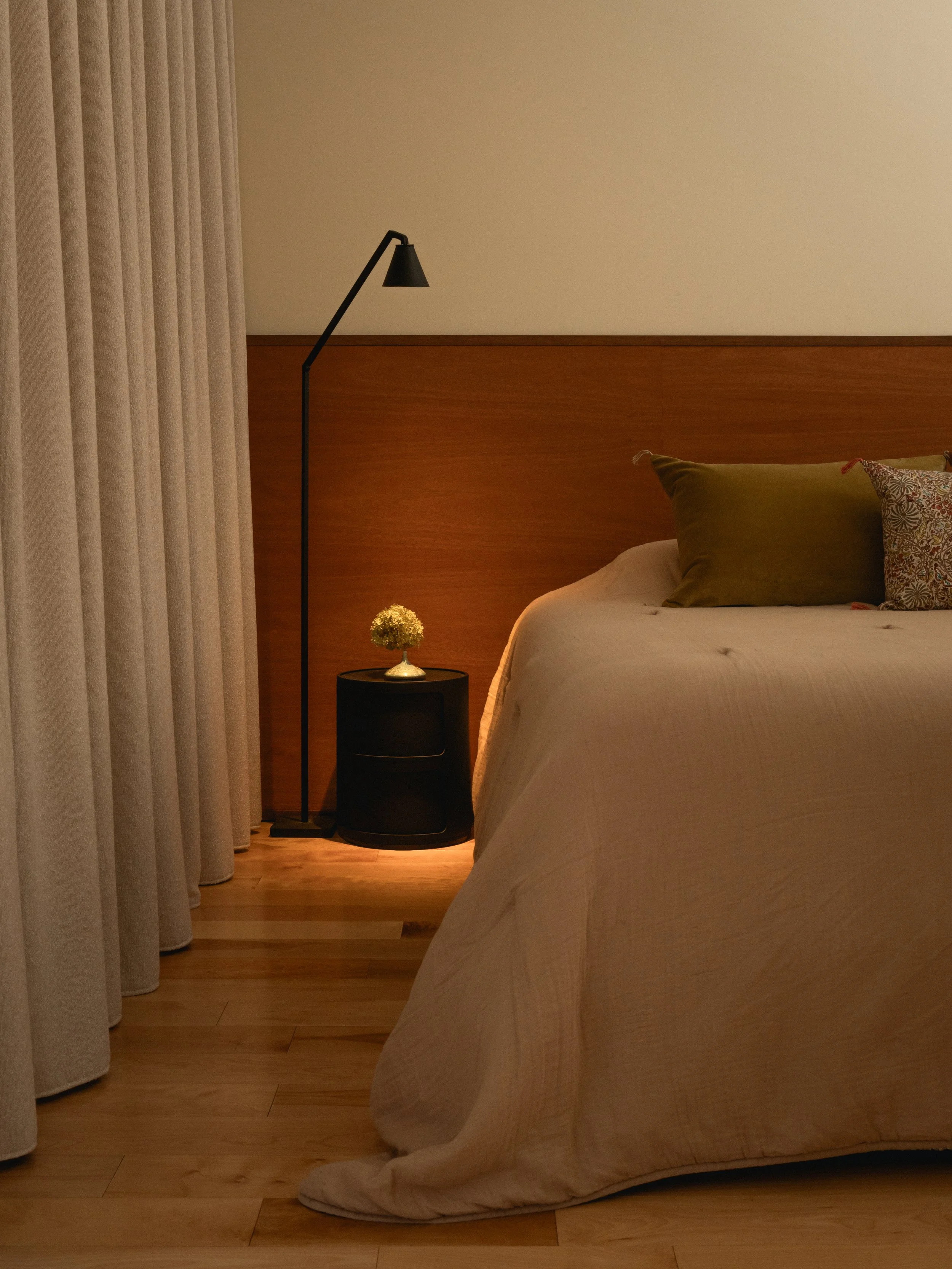Chambre avec un lit beige, une table d'appoint noire avec une petite plante dessus, une lampe à bras noir, et un volet ou rideau blanc cassé à gauche.