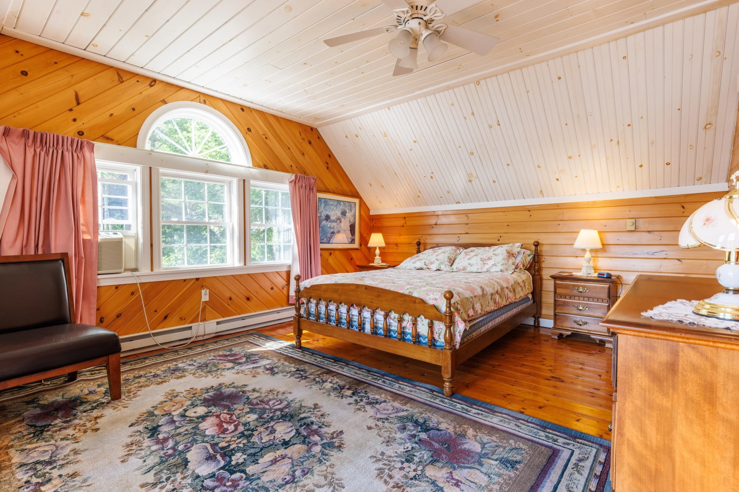 A bedroom with wooden panel walls and ceiling, a bed with floral bedding, a window with pink curtains, two lamps, a painting on the wall, and a colorful rug on the wooden floor.