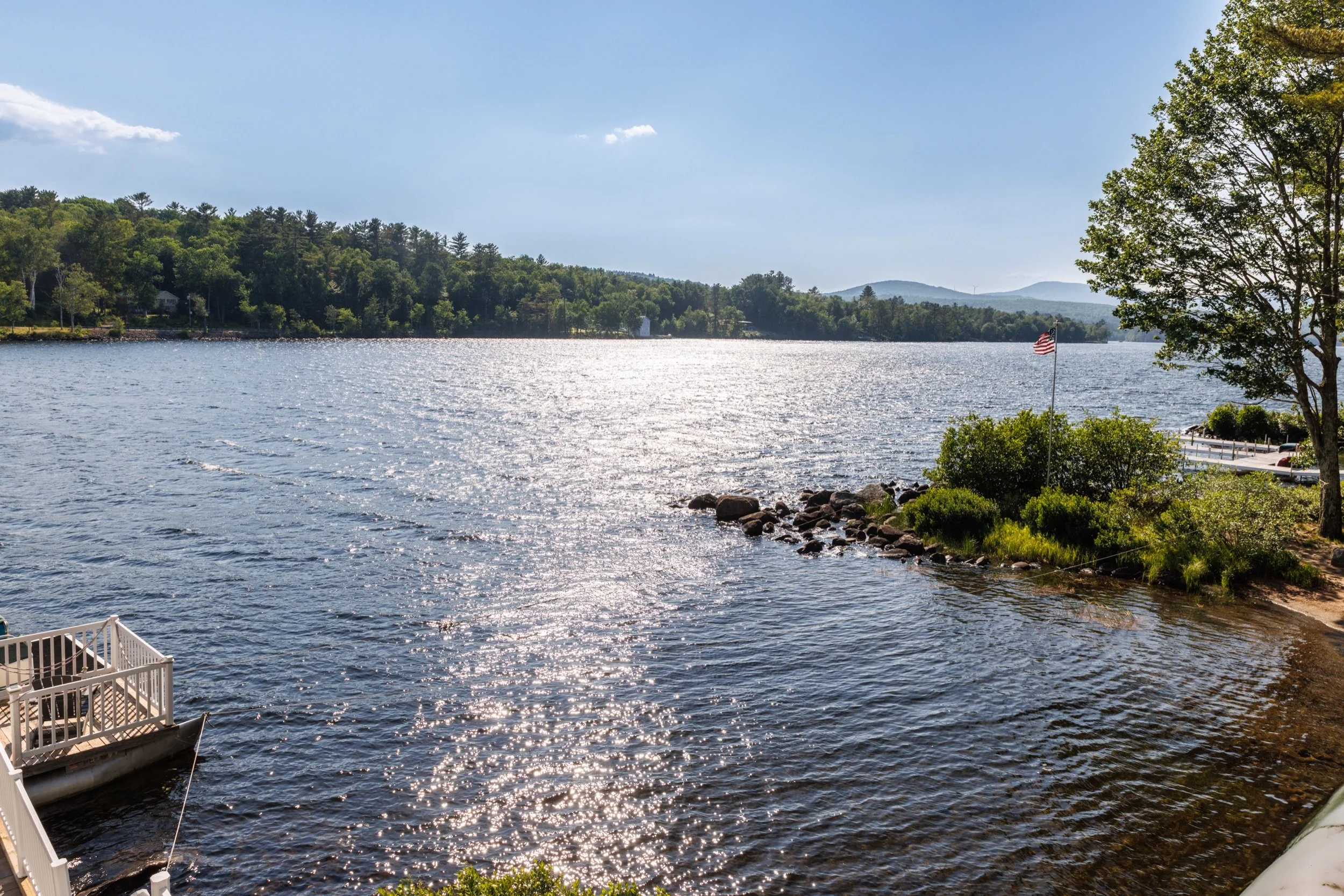 A peaceful lake scene with sunlight reflecting off the water, surrounded by green trees and distant hills, with American flag on a pole near the shoreline.
