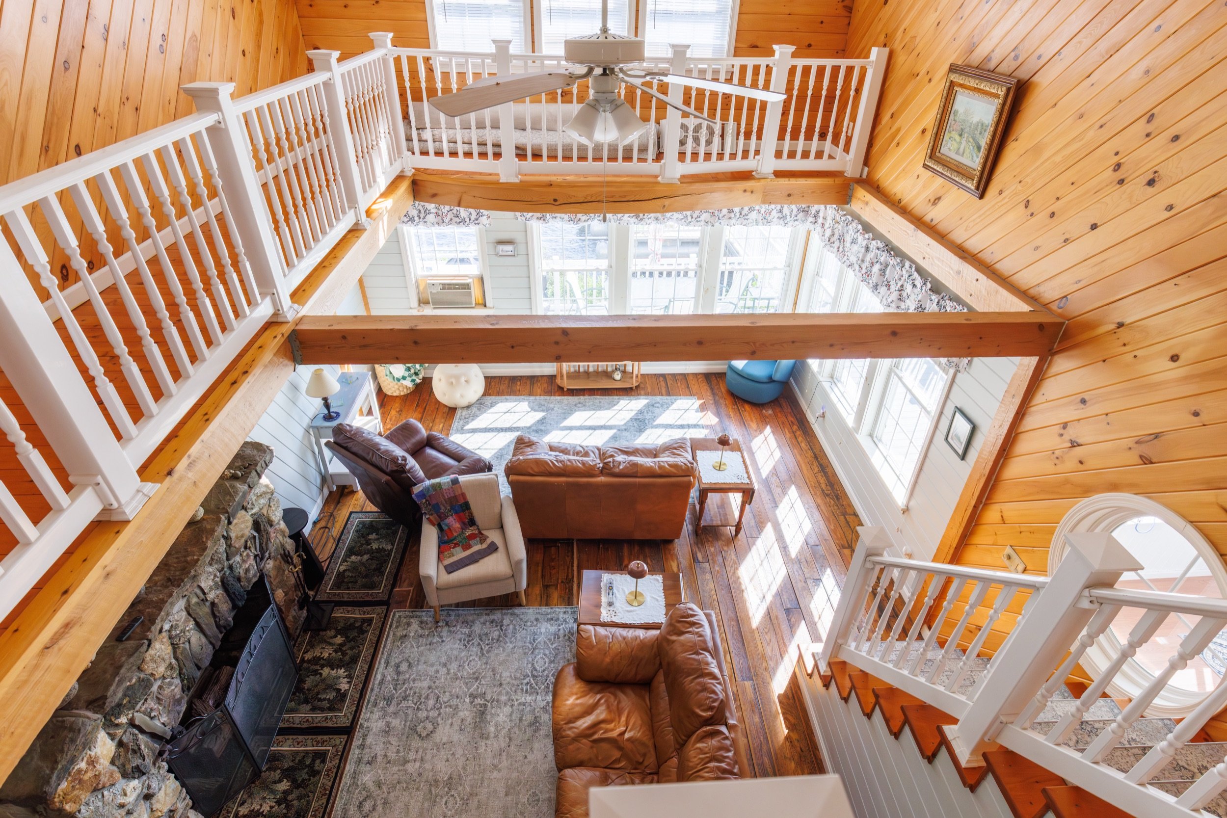 View from upstairs of a living room with wooden walls, a white railing balcony, large windows, and a roof fan. The living room has a brown leather sofa, armchairs, a fireplace, and a rug.