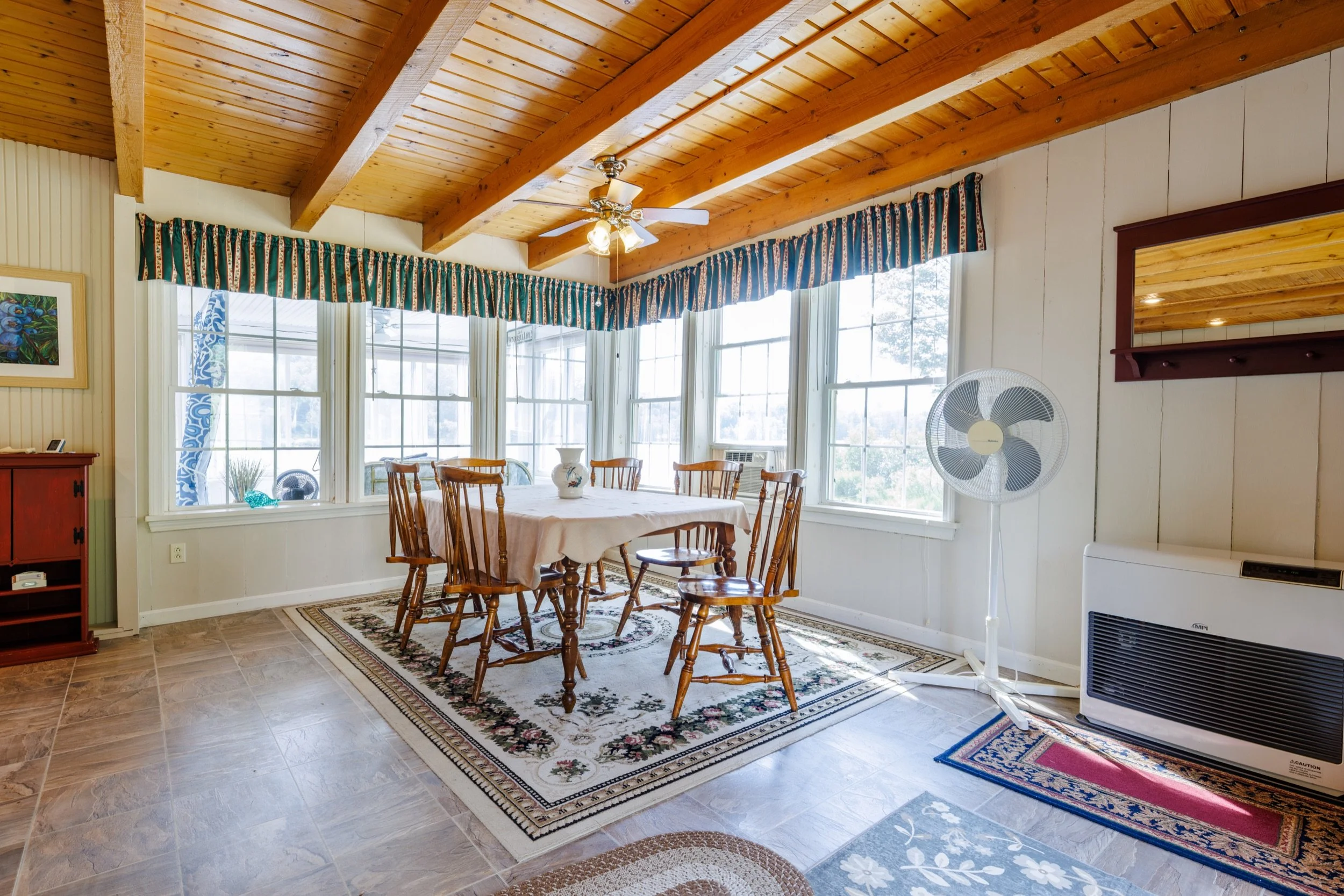 A dining room with wooden ceiling beams, large windows with patterned curtains, a wooden dining table with six chairs, a ceiling fan with lights, a decorative area rug, a bookshelf, a standing fan, and a heater.