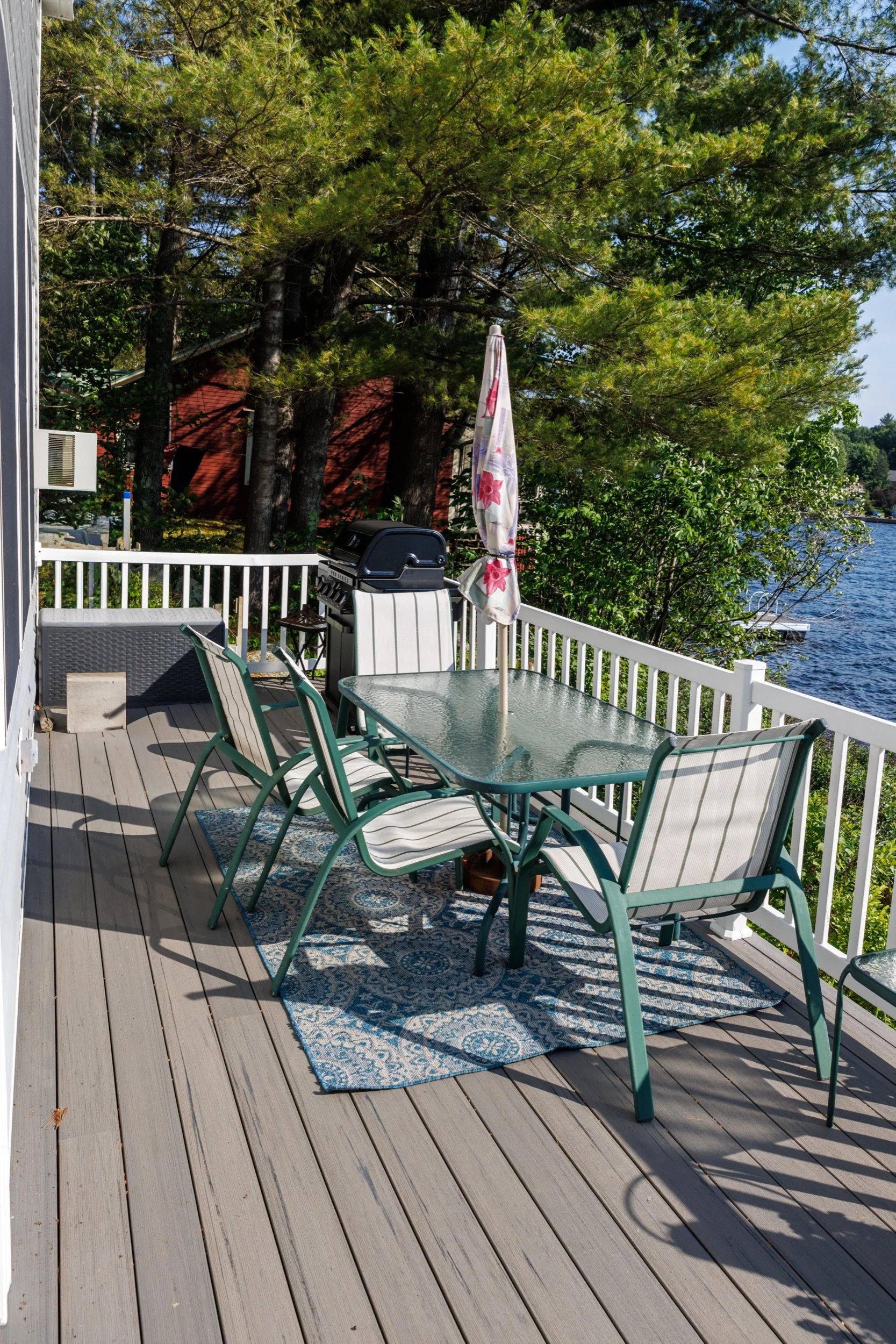 A backyard patio with a glass-top table and six metal chairs with striped cushions. There is a blue patterned outdoor rug under the table. A white railing surrounds the patio, which overlooks a body of water. A covered barbecue grill is next to the t