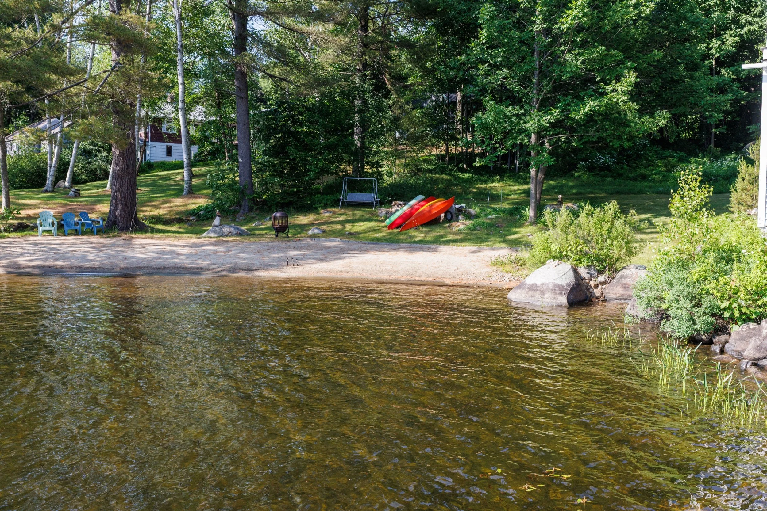A lakeside backyard with a sandy beach, green grass, tall trees, and a small house partially visible behind the trees. There are colorful kayaks, Adirondack chairs, a swing, and a small fire pit near the shore.