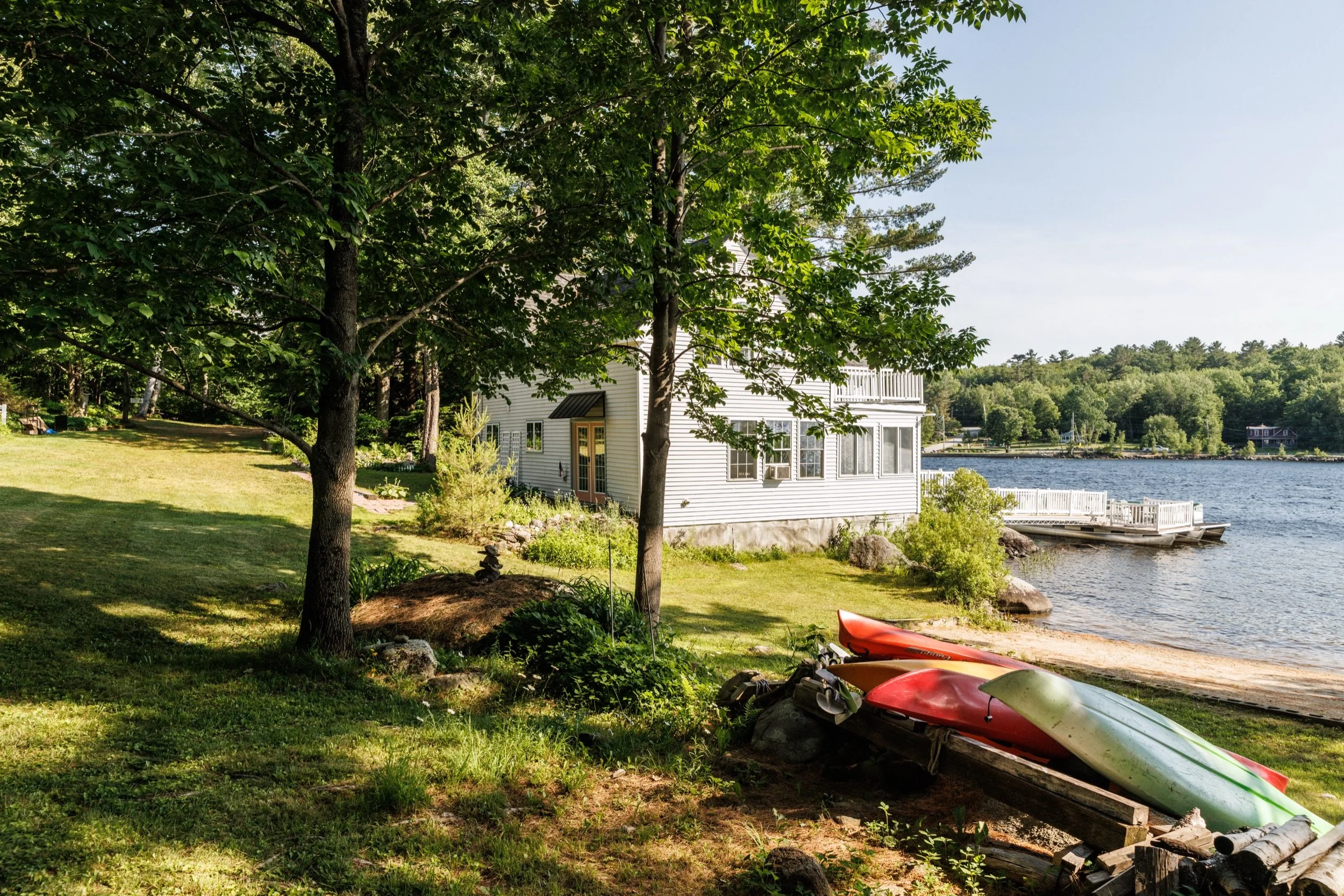 View of a lakeside yard with a white house, green trees, and two kayaks on the grass near the water.