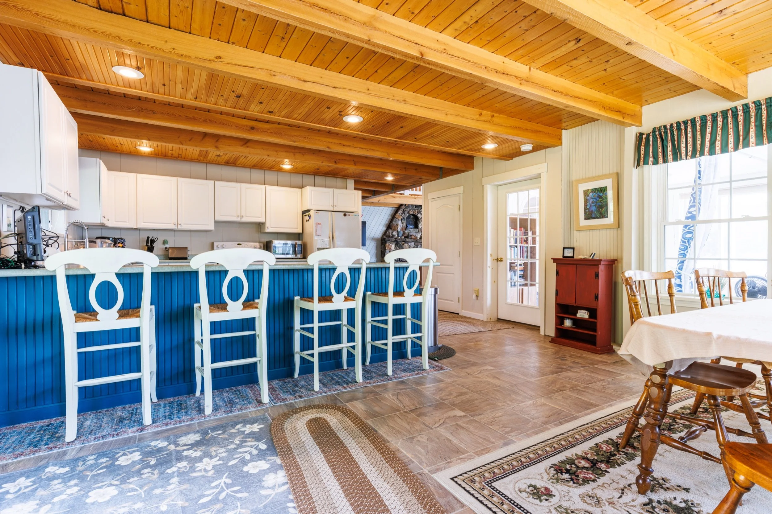 Interior view of a kitchen and dining area with a wooden ceiling, white cabinets, a blue kitchen island with bar stools, a fireplace, and windows with green curtains.