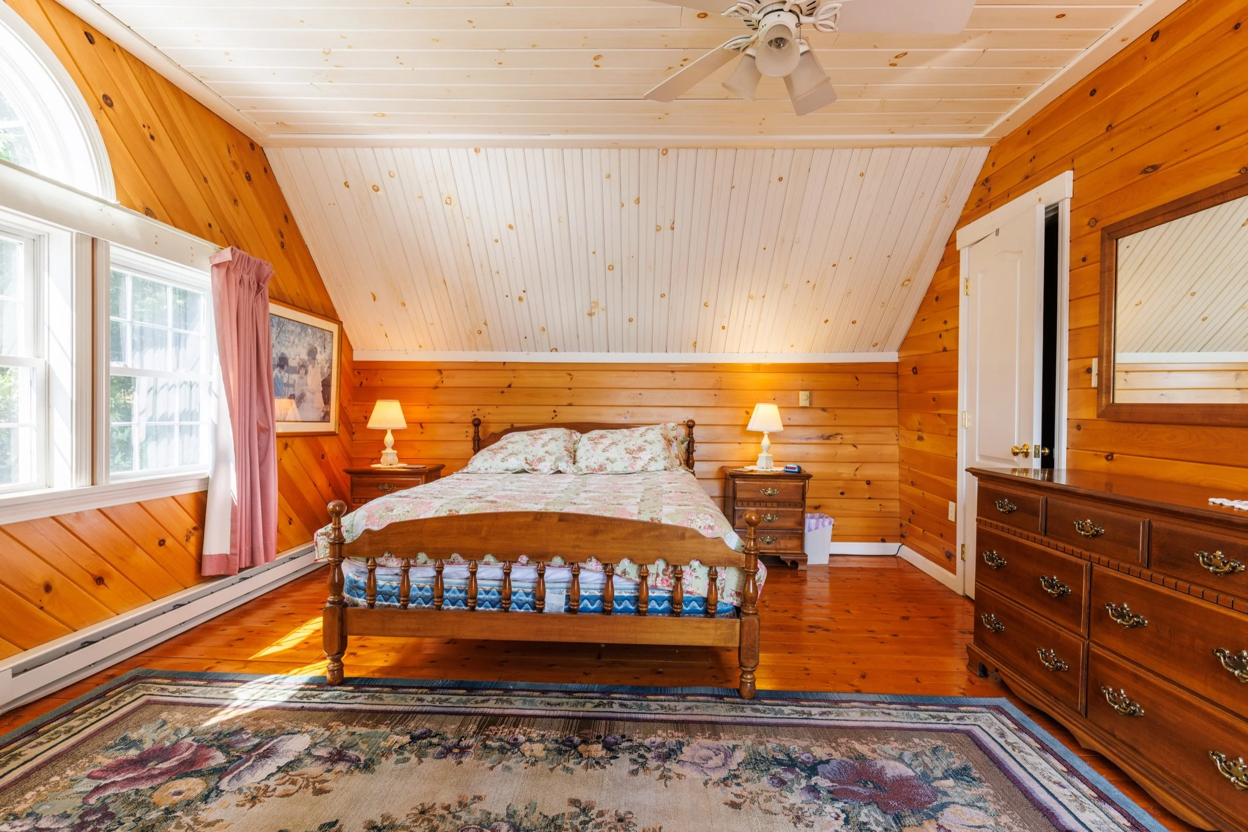A cozy bedroom with wood-paneled walls and ceiling, a wooden bed with floral bedding, two bedside lamps, a window with pink curtains, and a wooden dresser with a mirror.