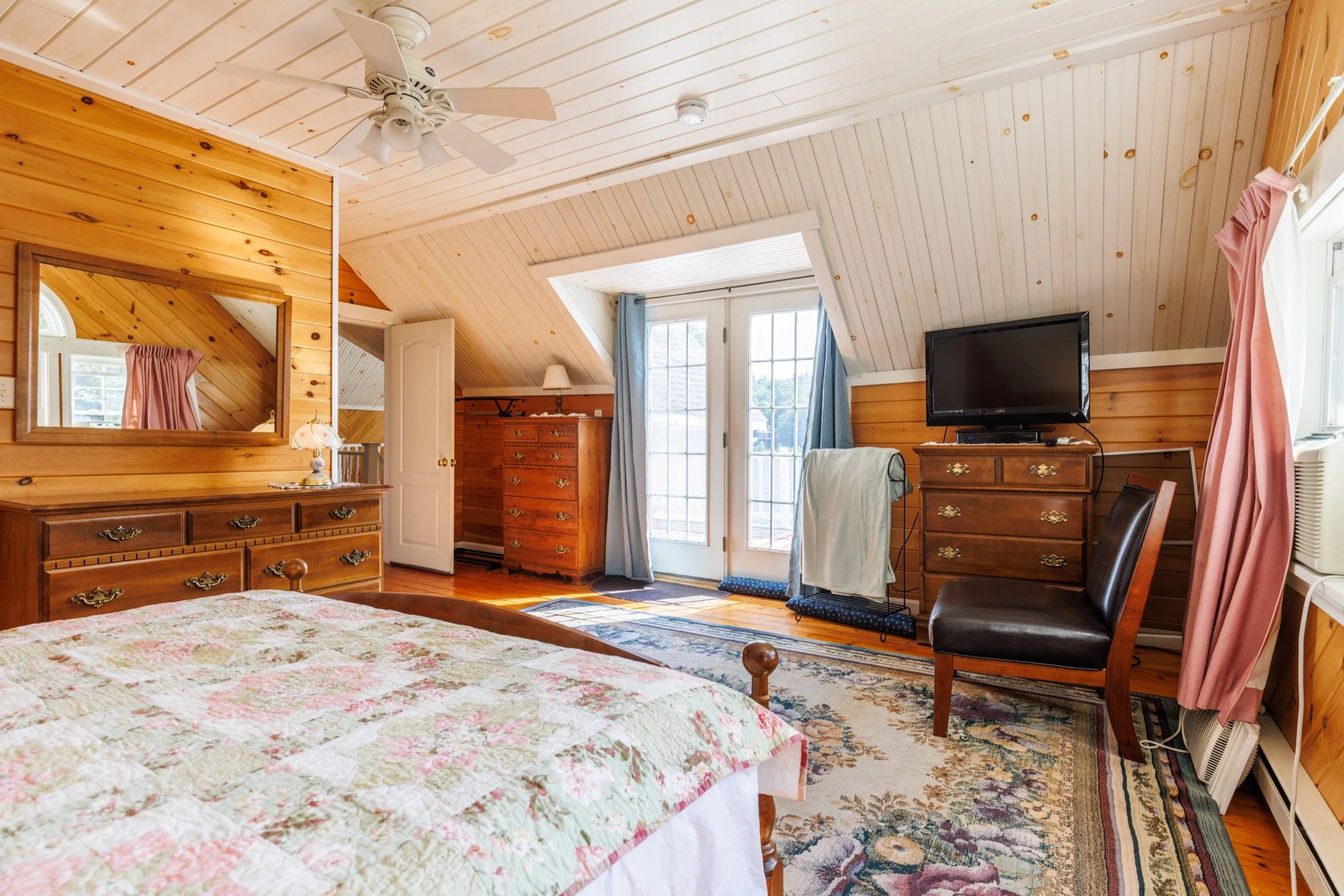 A cozy bedroom with wood-paneled walls and ceiling, a bed with floral bedding, a dresser with a lamp, a TV on a stand, and windows with blue and pink curtains.