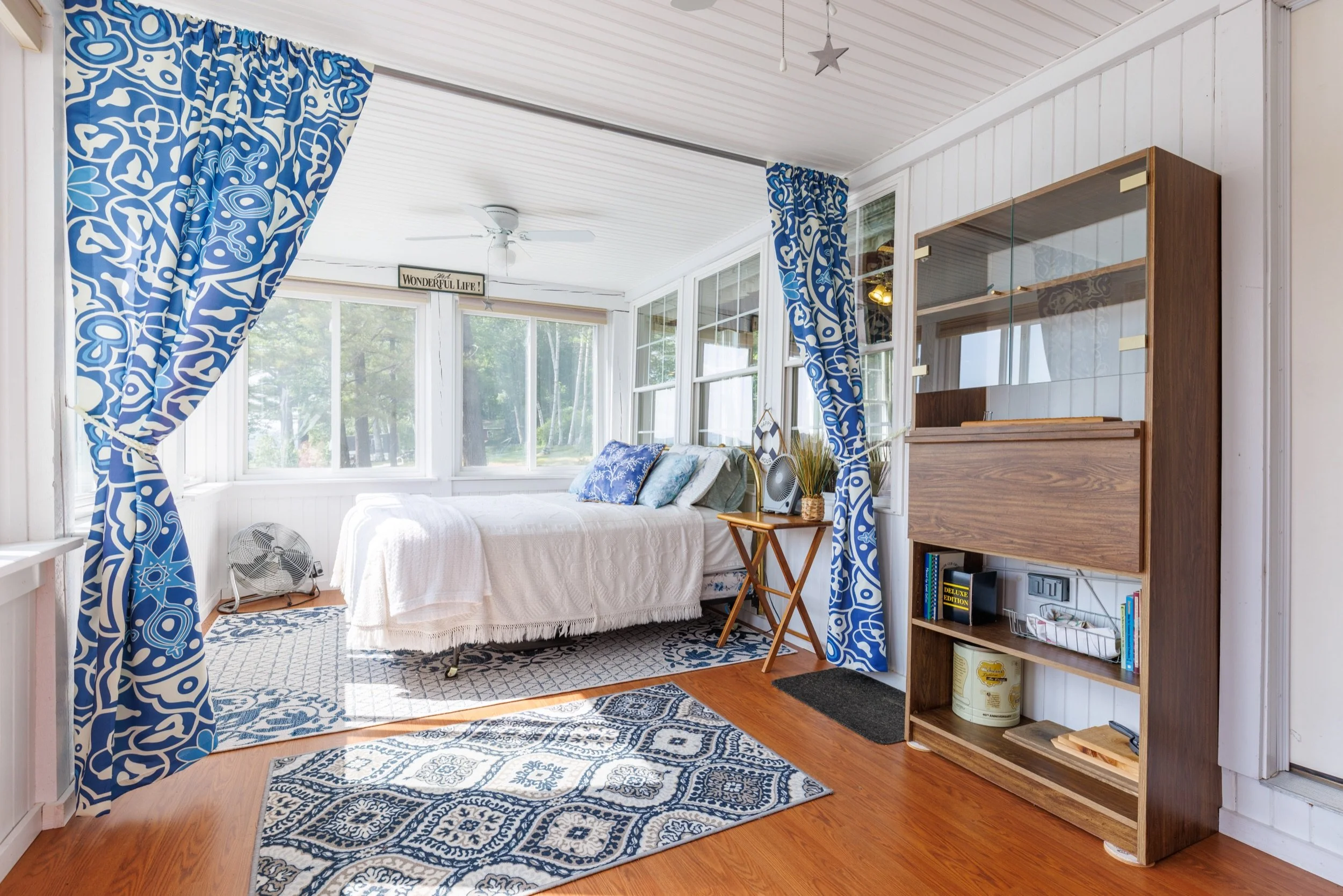 Sunroom with large windows, blue patterned curtains, a white bed with pillows, a wooden shelving unit, ceiling fan, and a patterned area rug.