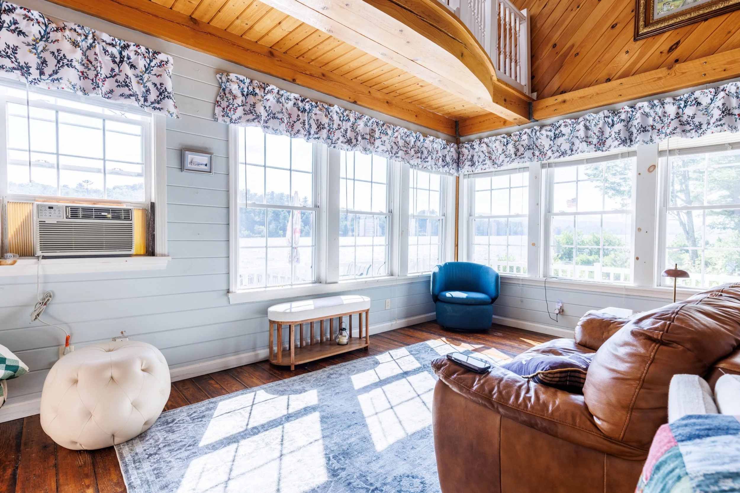 A cozy living room with large windows letting in sunlight, wooden ceilings, and a mix of furniture including a blue armchair, a leather sofa, and a white tufted ottoman. There's a small wooden side table and a patterned area rug on the hardwood floor