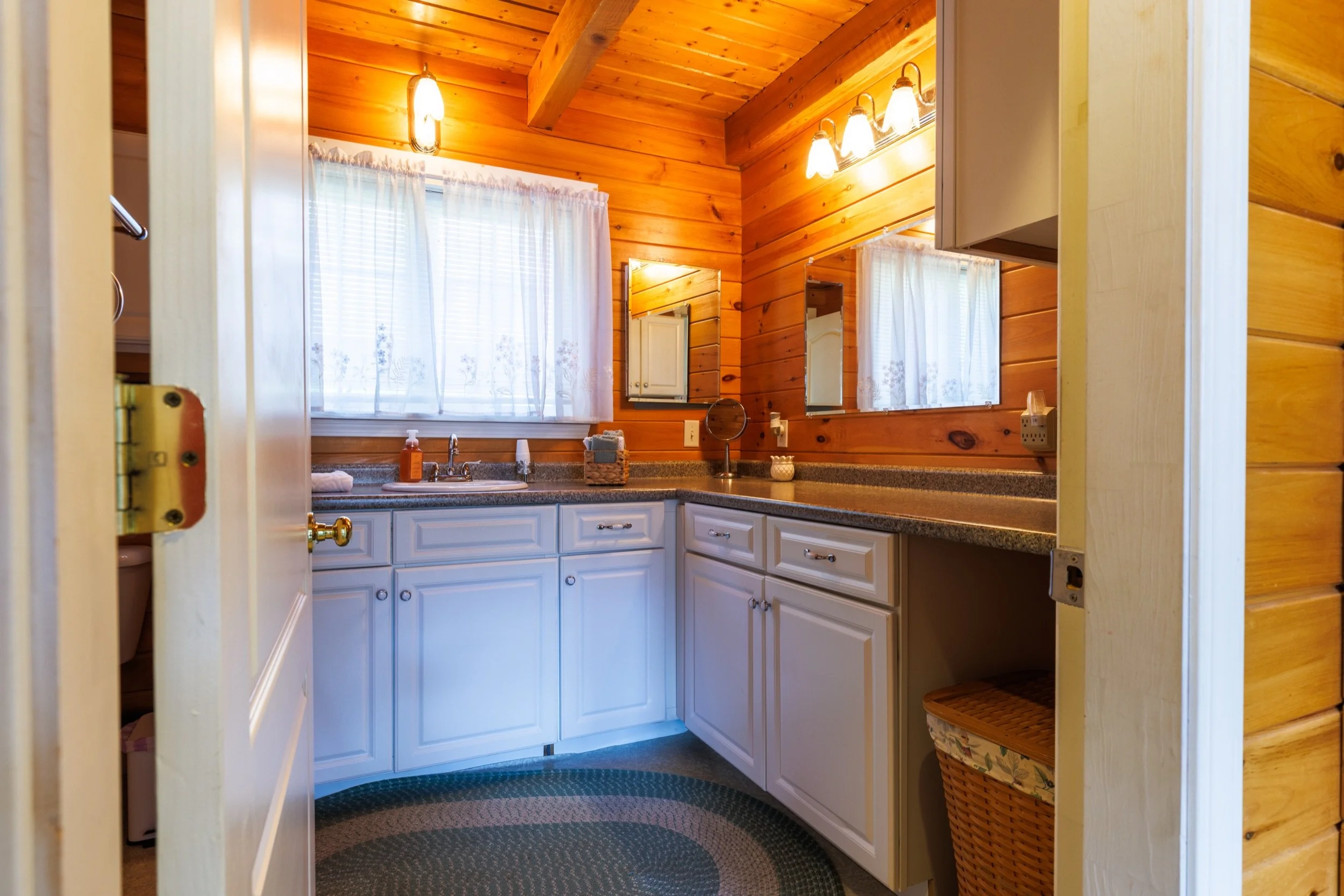 View of a cozy bathroom with wooden-paneled walls and ceiling. It features a double sink vanity with white cabinets, a woven basket on the right, and two mirrors above the sinks. The bathroom is lit by four wall-mounted light fixtures, and sheer curt