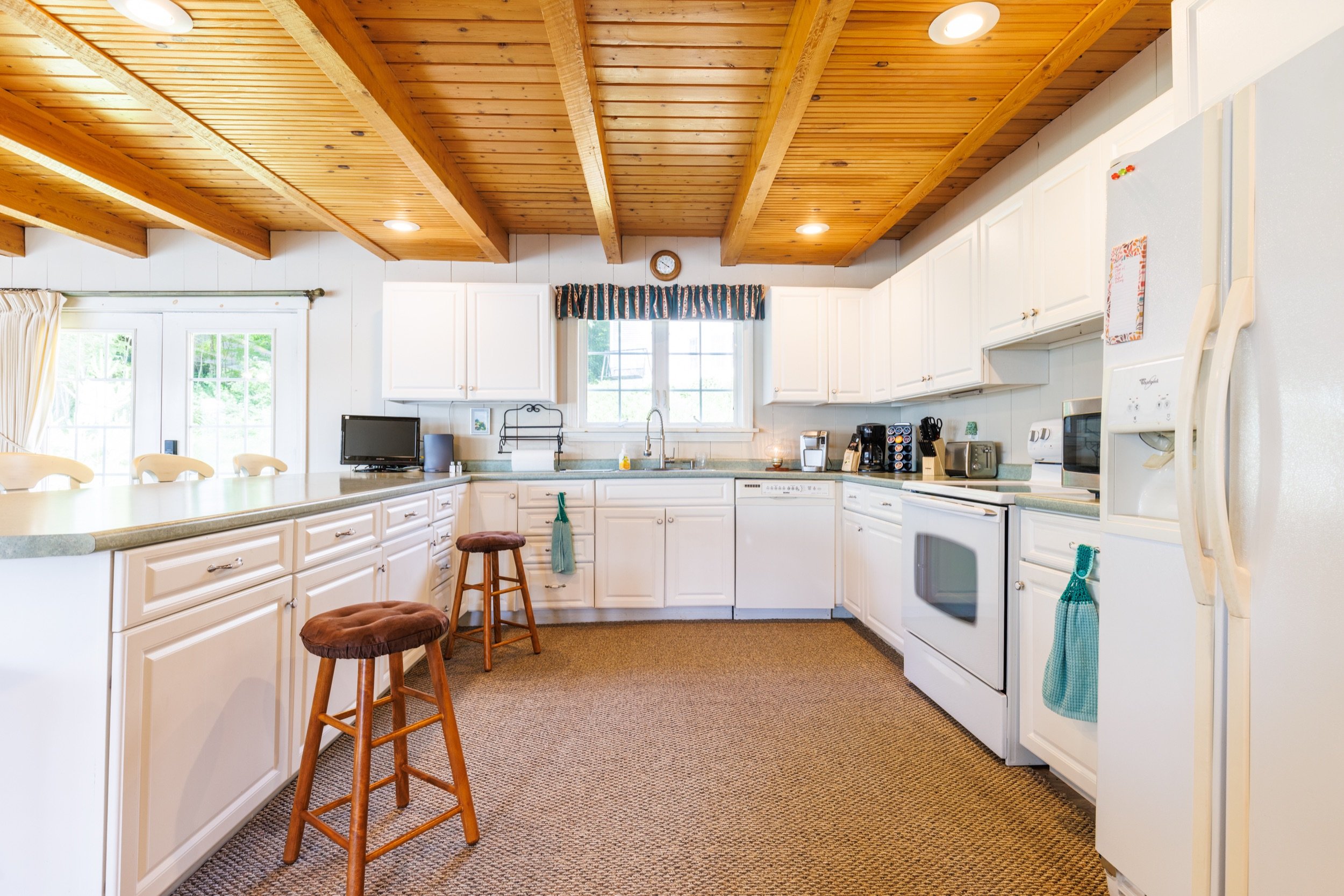 A kitchen with white cabinets, a wooden ceiling with exposed beams, and a window with a striped valance. The counter has a small TV, a coffee maker, and other appliances. There are two brown stools and a brown carpet on the floor.