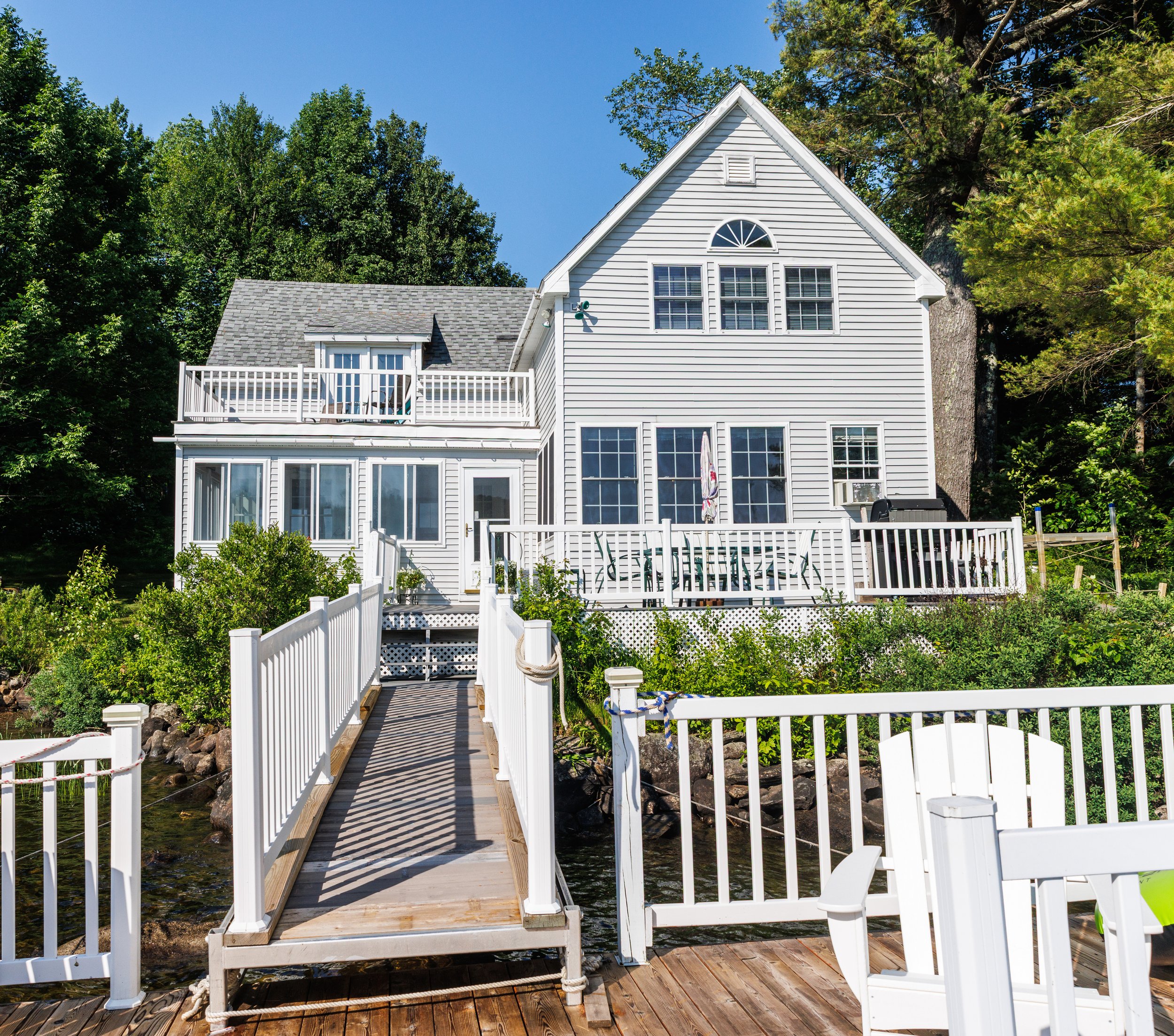 White house with multiple windows and a balcony, surrounded by green trees under a clear blue sky, with a wooden dock and white fence leading to water.