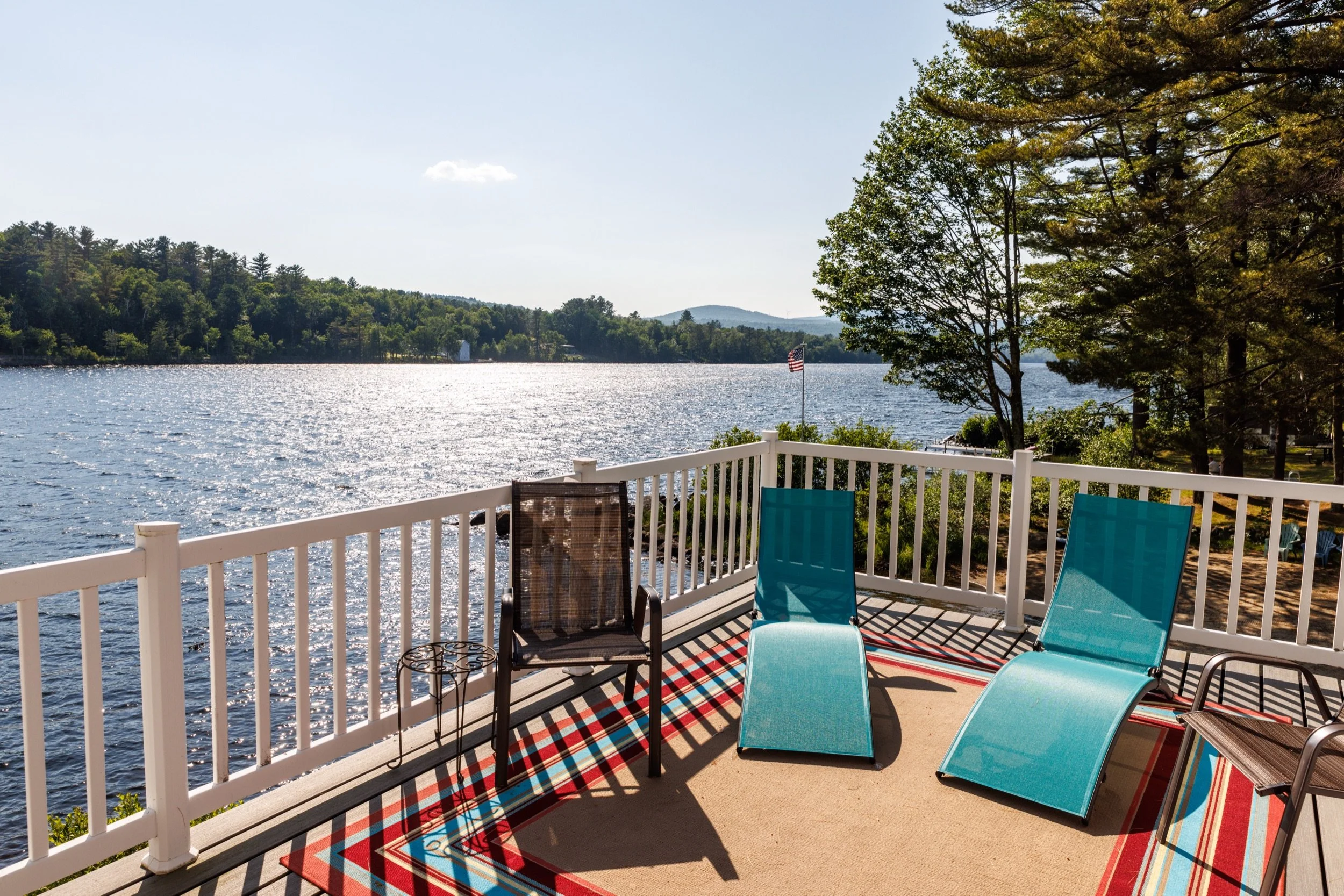 View of a lakeside from a outdoor balcony with a white railing, featuring several lounge chairs in blue and brown, a patterned rug, and an American flag on a flagpole, with a body of water, green trees, and hills in the background.