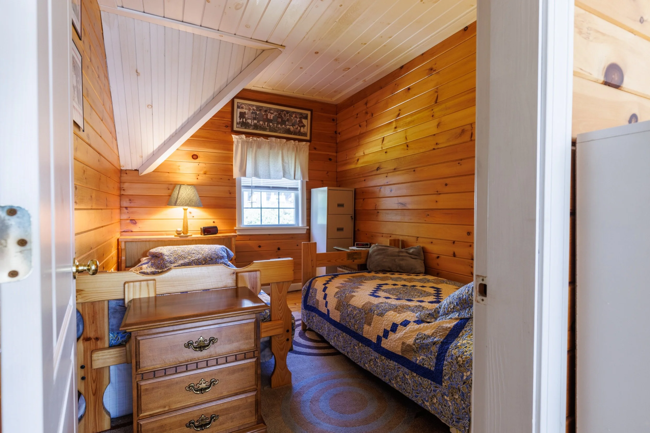 A cozy bedroom with wood-paneled walls and ceiling. It has a small window with a white curtain, a bed with a quilted cover, a bedside table with a lamp, and a framed picture on the wall.