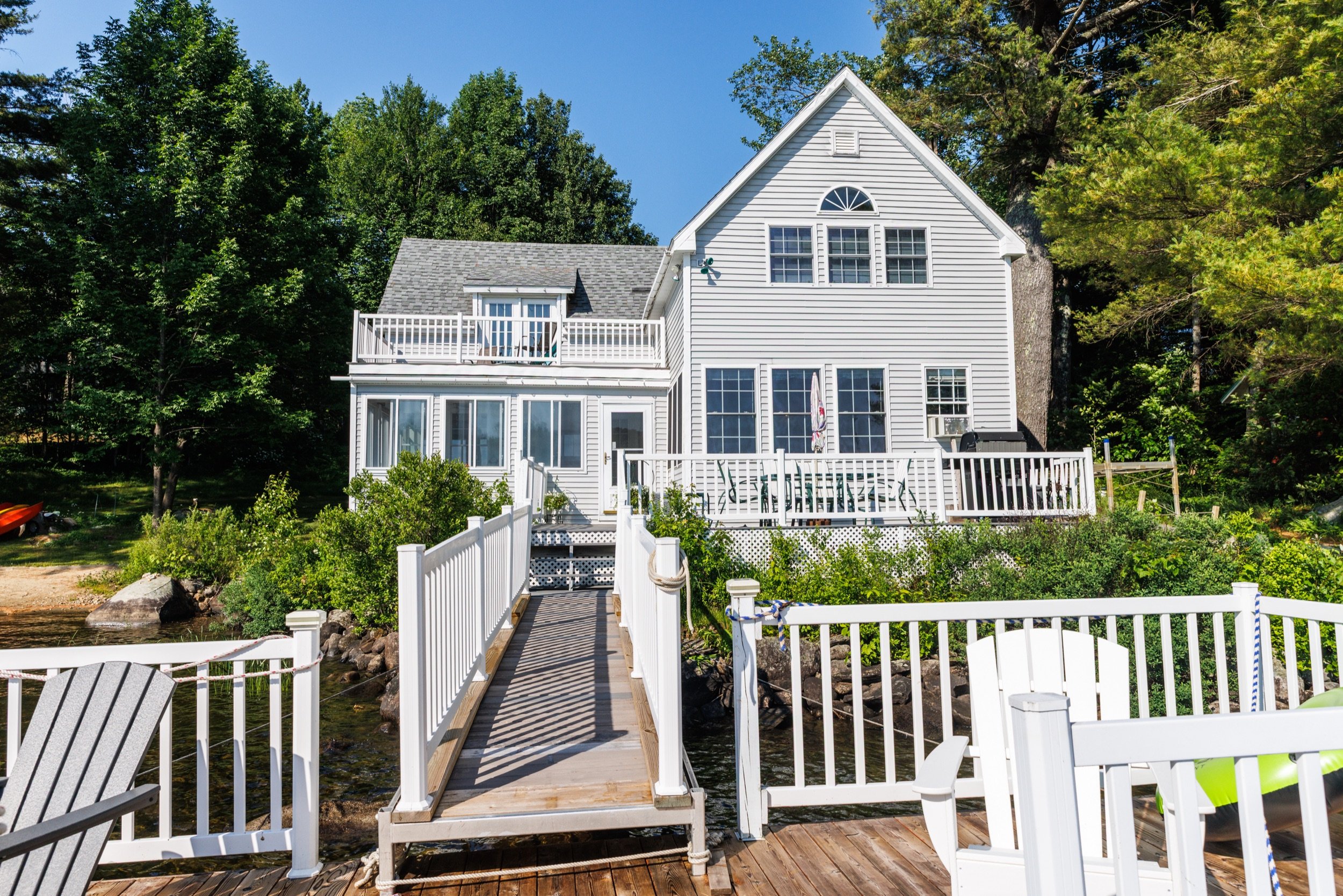 White house with multiple windows and a balcony surrounded by trees, with a dock and benches in the foreground.