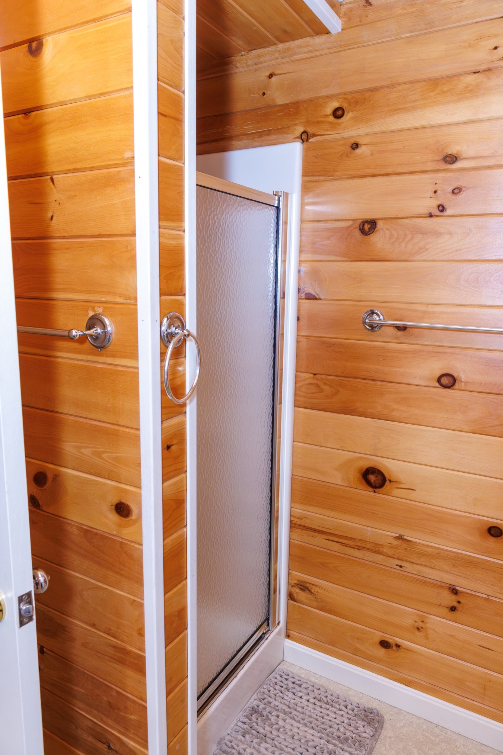 Inside a bathroom with wood-paneled walls, a glass shower door, a towel bar, and a small textured rug on the floor.