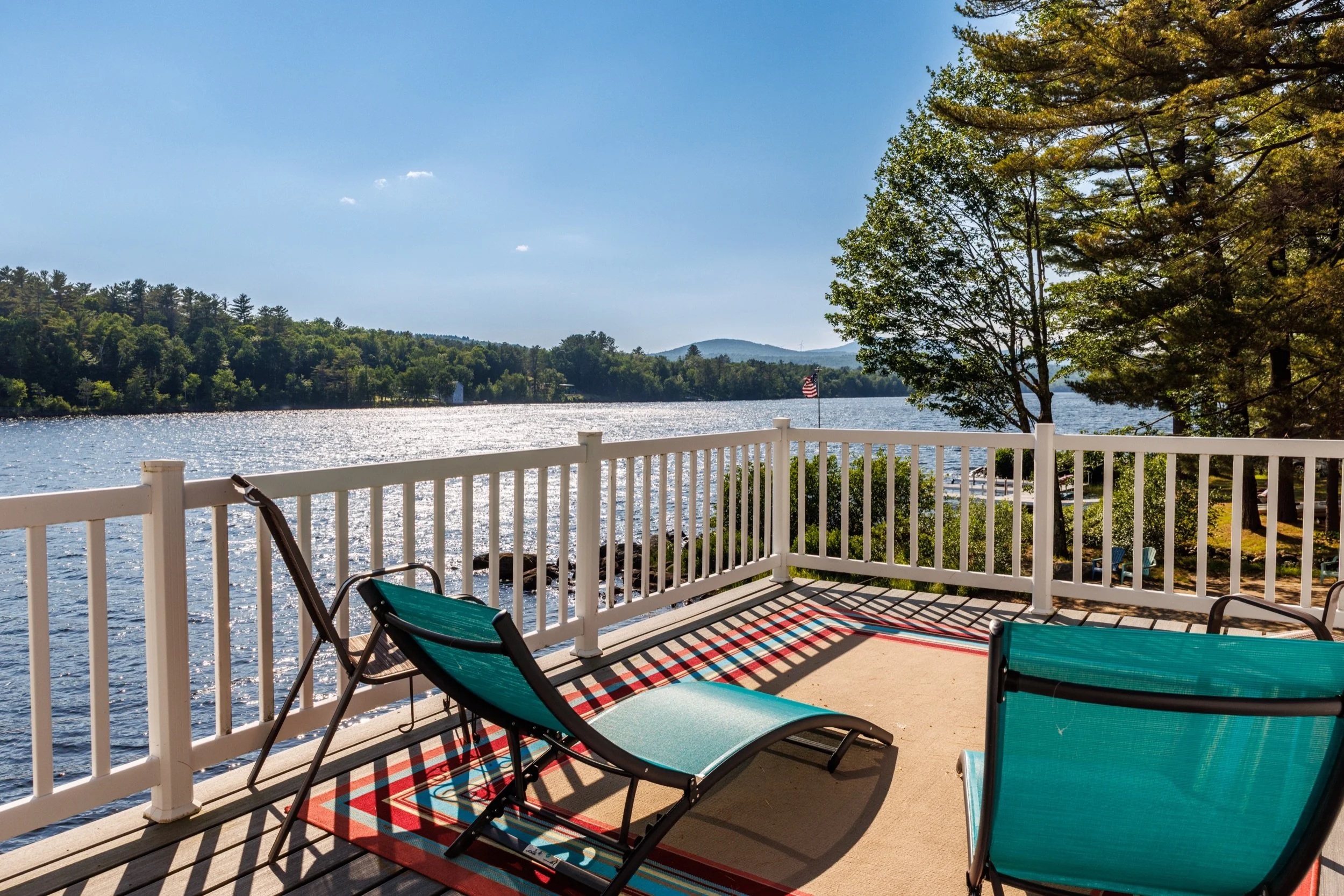 View from a porch overlooking a lake with trees, hills, and a blue sky. The porch has chairs and a white railing.