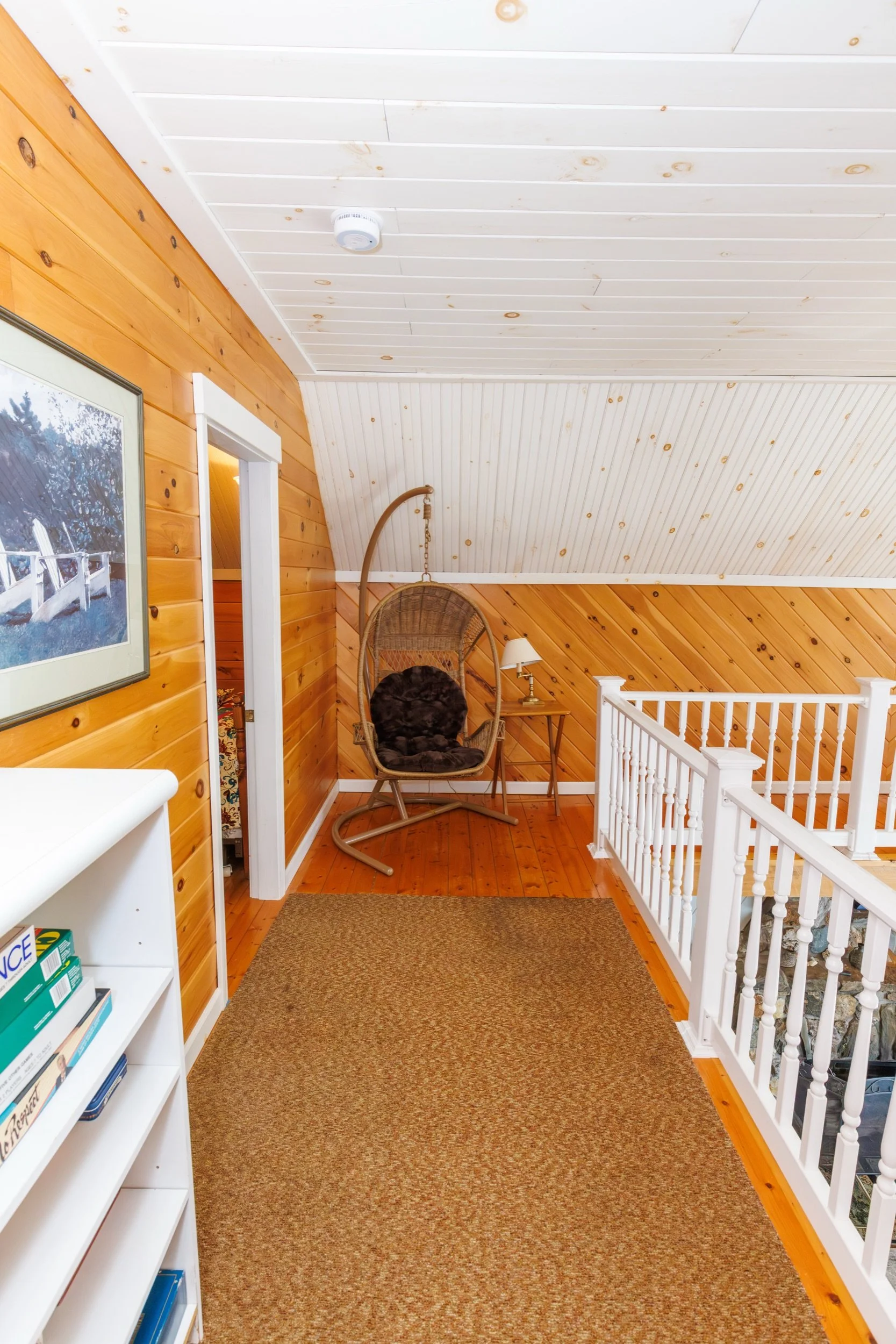 A cozy, enclosed upstairs porch with wood-paneled walls and ceiling. It features a hanging papasan chair, a small side table, a lamp, a framed picture on the wall, a storage shelf with books or magazines, and a tan rug on the wooden floor.