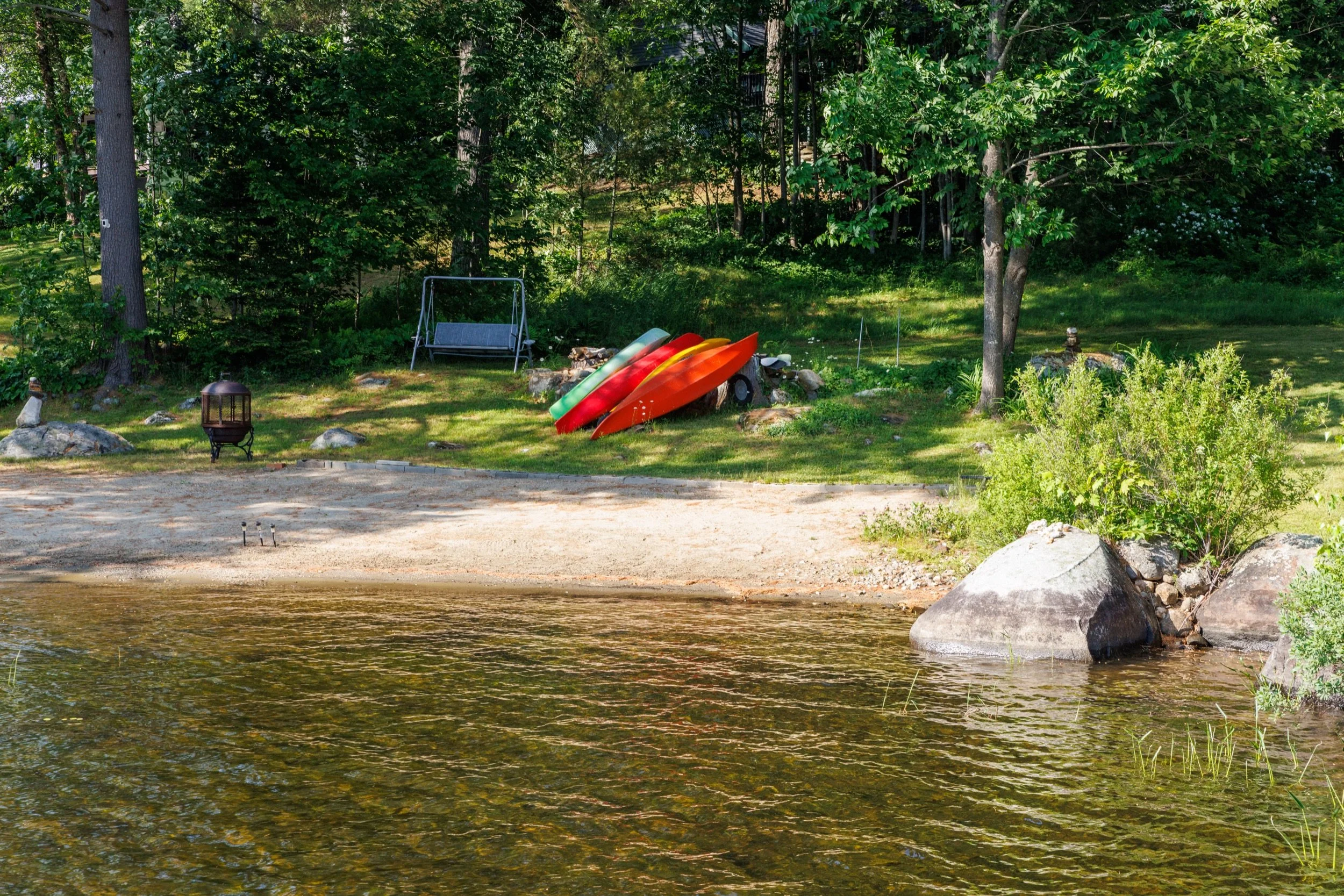 A lakeside yard with a grassy area, trees, a swing, several colorful kayaks stacked on a log, and rocks near the shoreline.