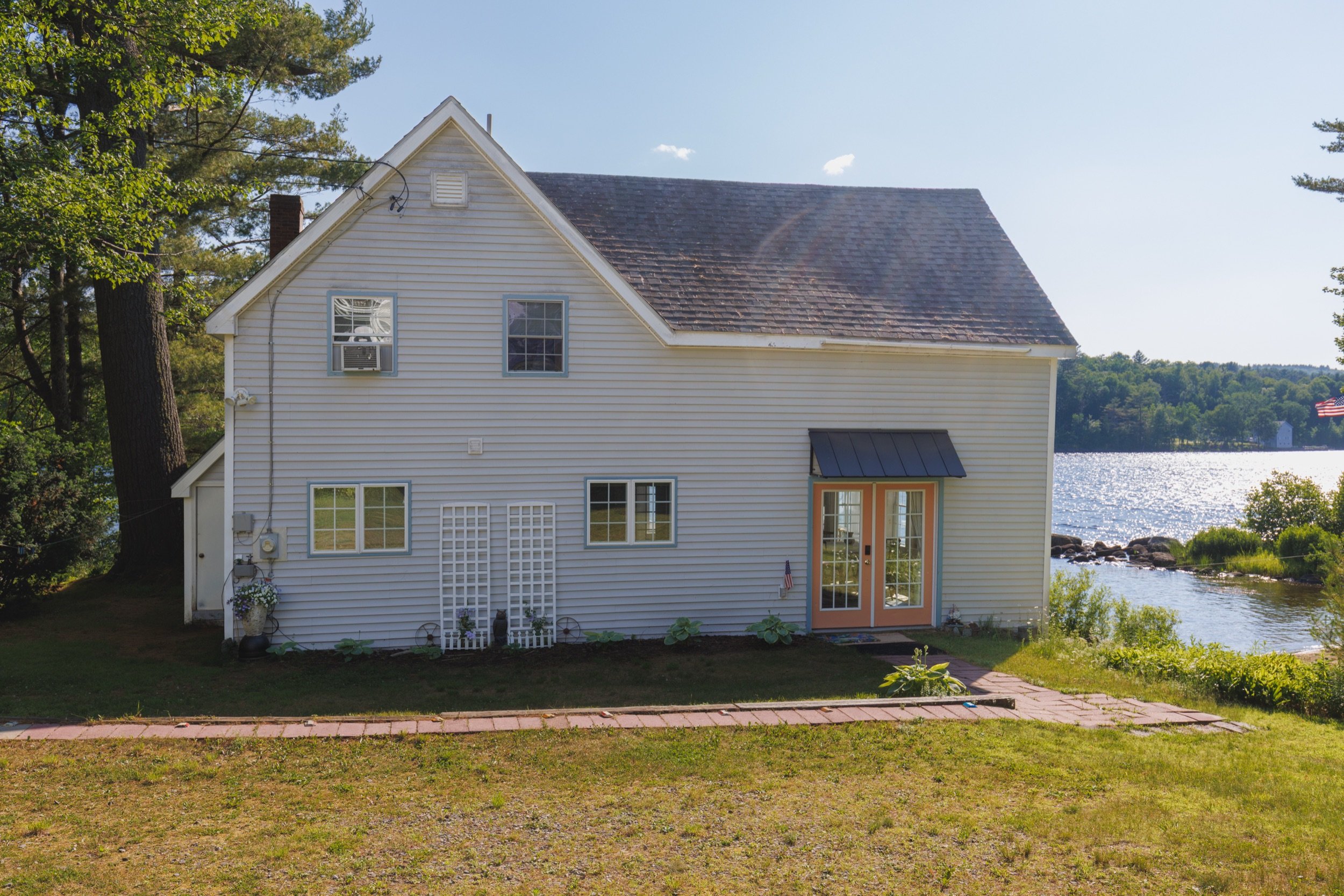 White house with a gable roof, situated near a body of water, with trees and a flag visible in the background.